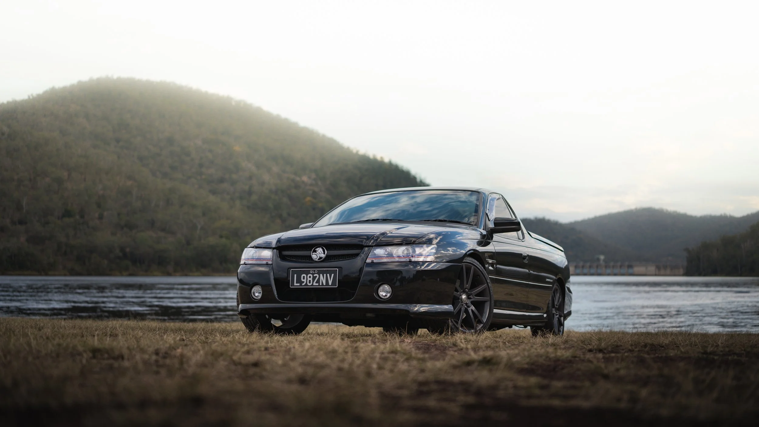A black Holden Monaro car parked on grass near a body of water with trees and hills in the background.