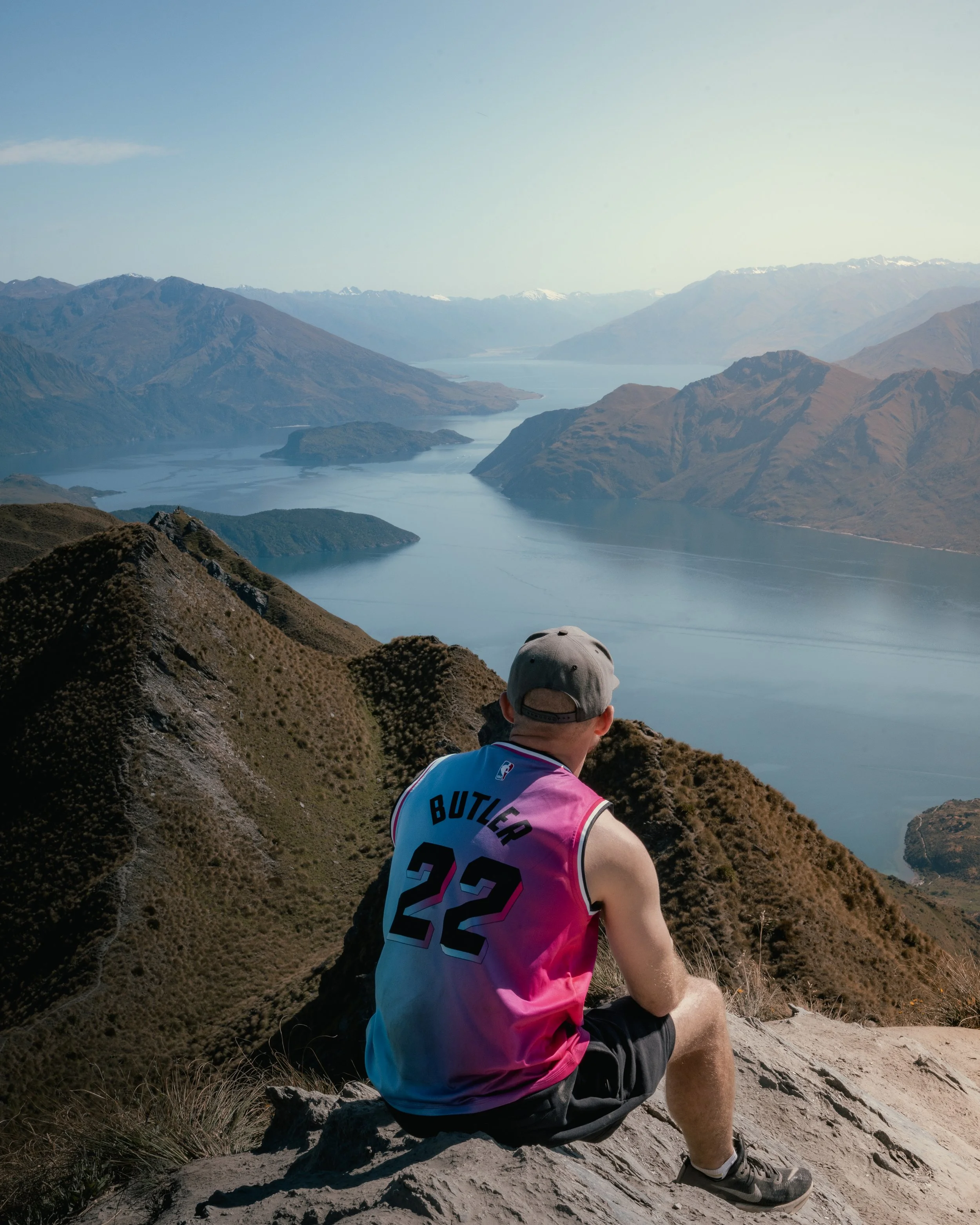A man sitting on a mountain trail overlooking a scenic lake and mountain range with snow-capped peaks in the distance.