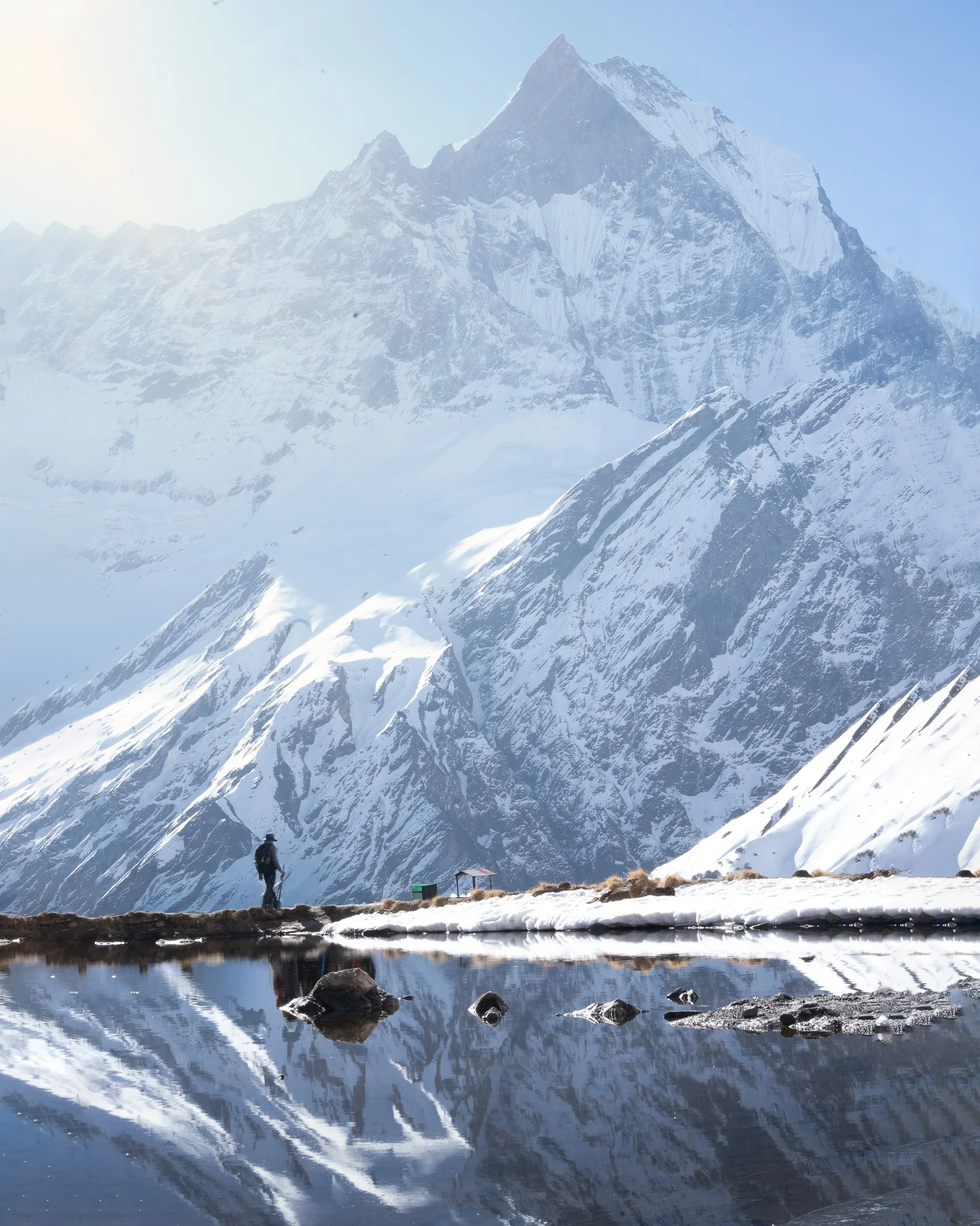 A snow-covered mountain range with a person walking near a small body of water, mountains reflected in the water, blue sky, and a small shelter or structure near the person.