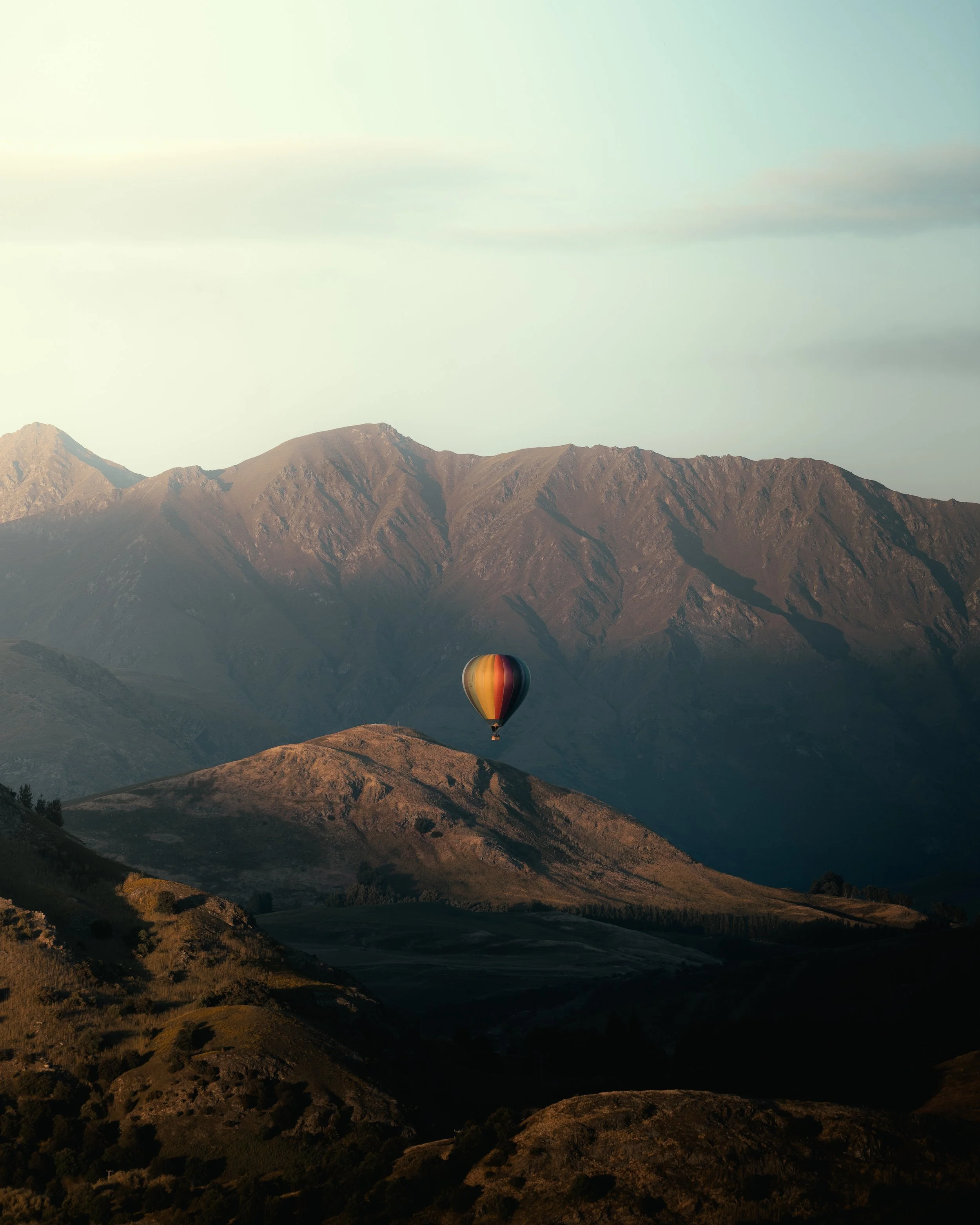 A hot air balloon with vertical stripes of red, yellow, and black floating above a mountain landscape during dusk or dawn.
