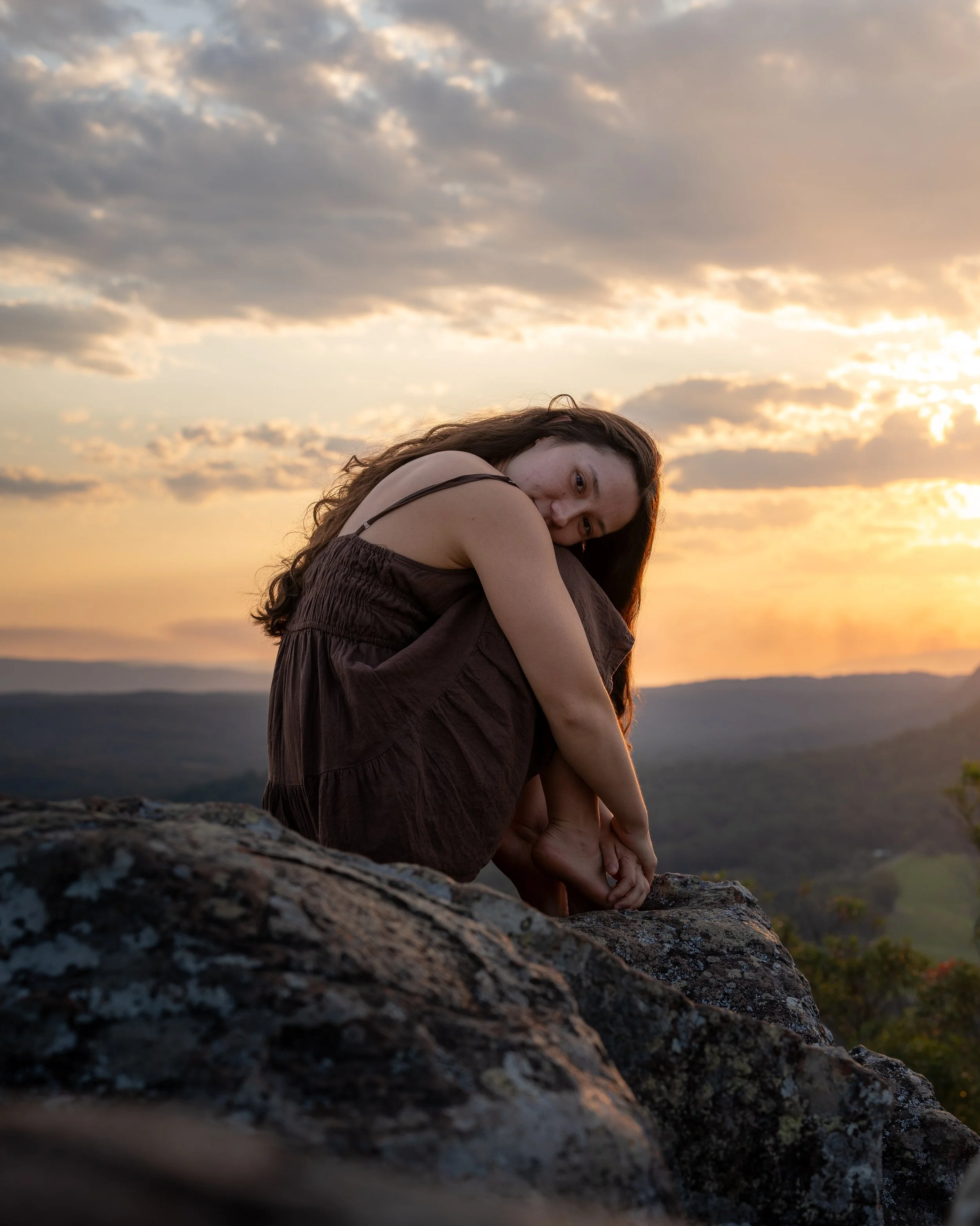 A woman with long brown hair sitting on a mountain rock at sunset, with her arms wrapped around her legs, looking at the camera, with a cloudy sky and distant mountains in the background.