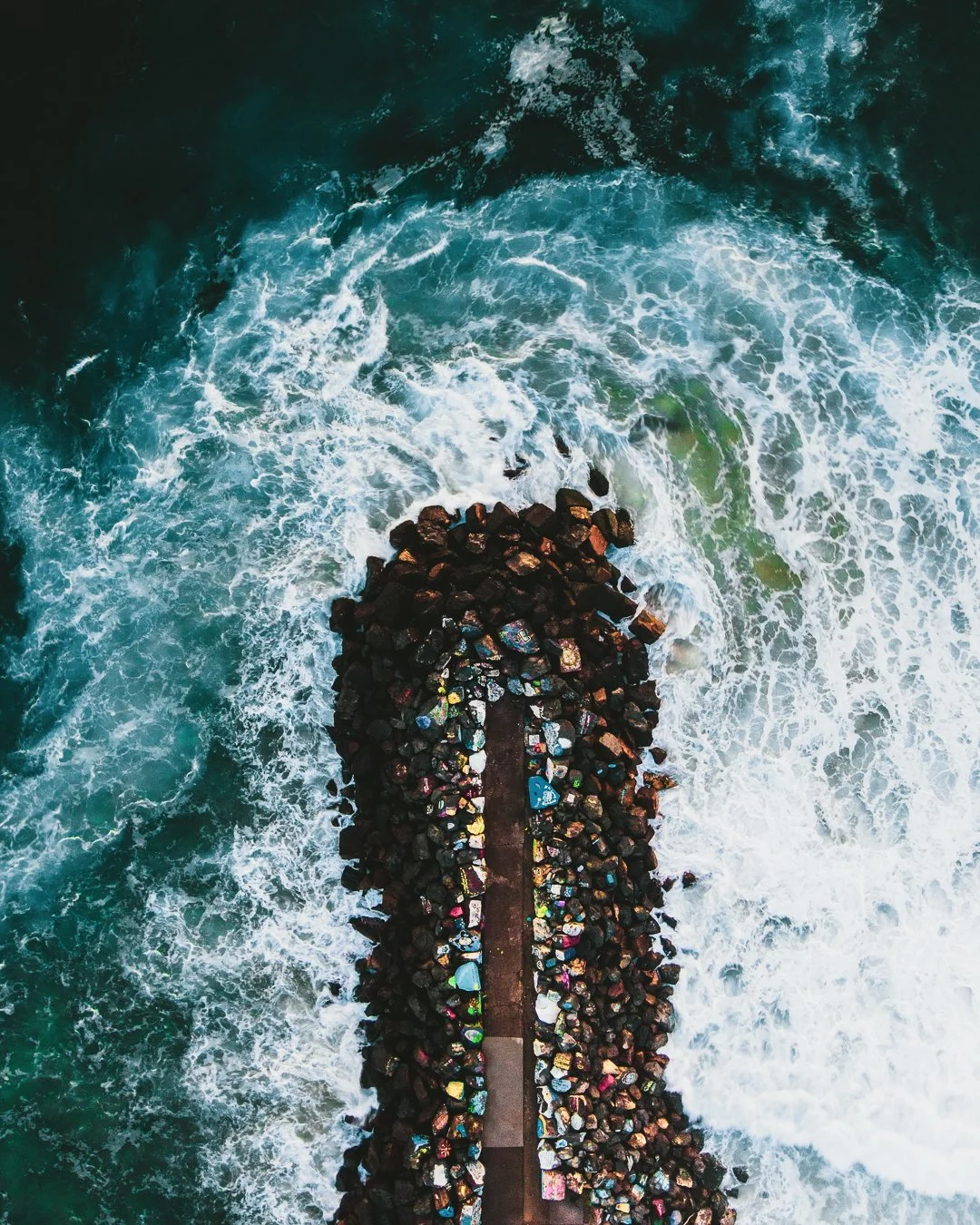 An aerial view of a rocky jetty extending into the ocean with waves crashing against it.