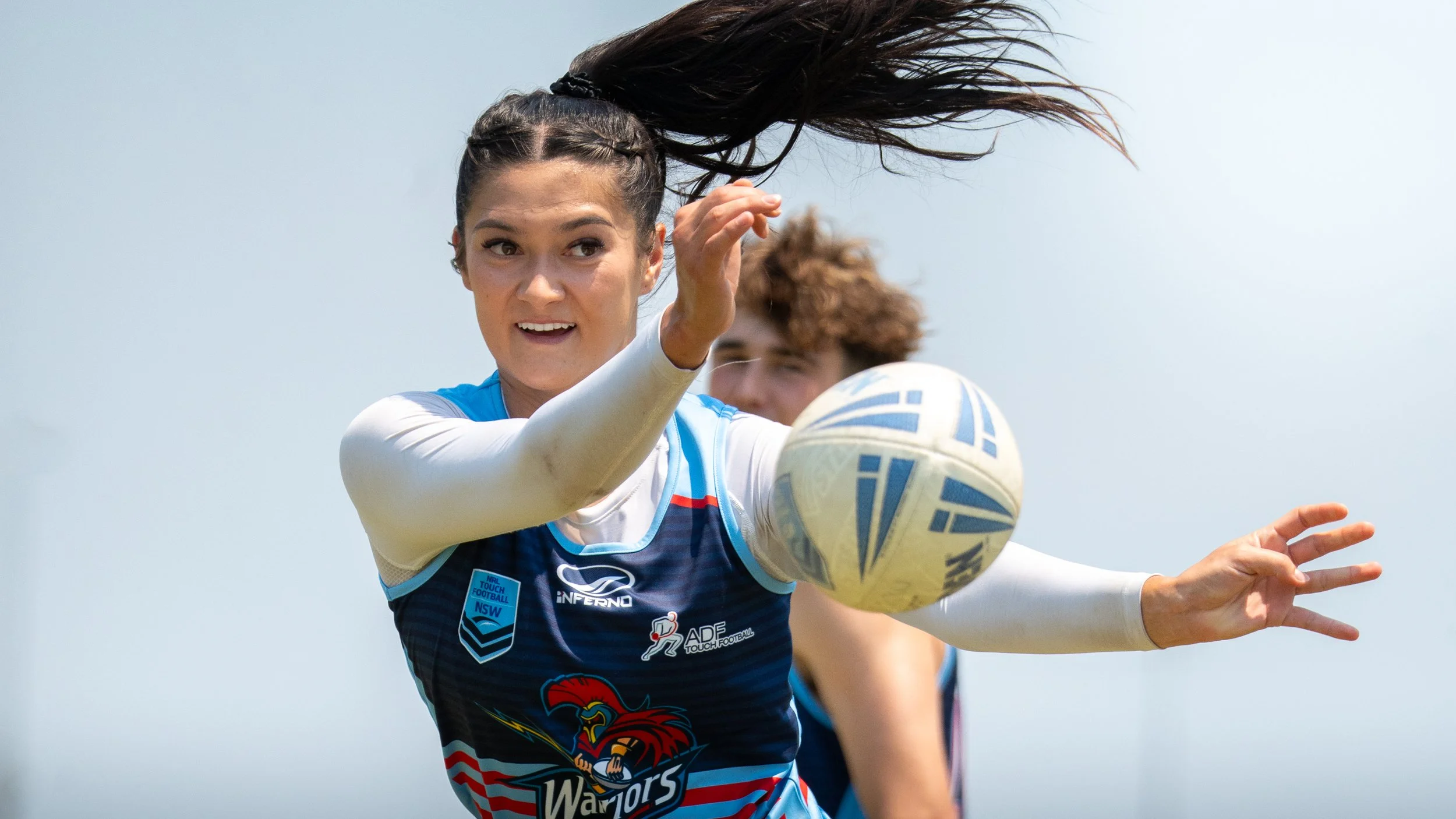 A female rugby player in a blue and white uniform throws a rugby ball during a game, with a focused expression and her long hair flying.