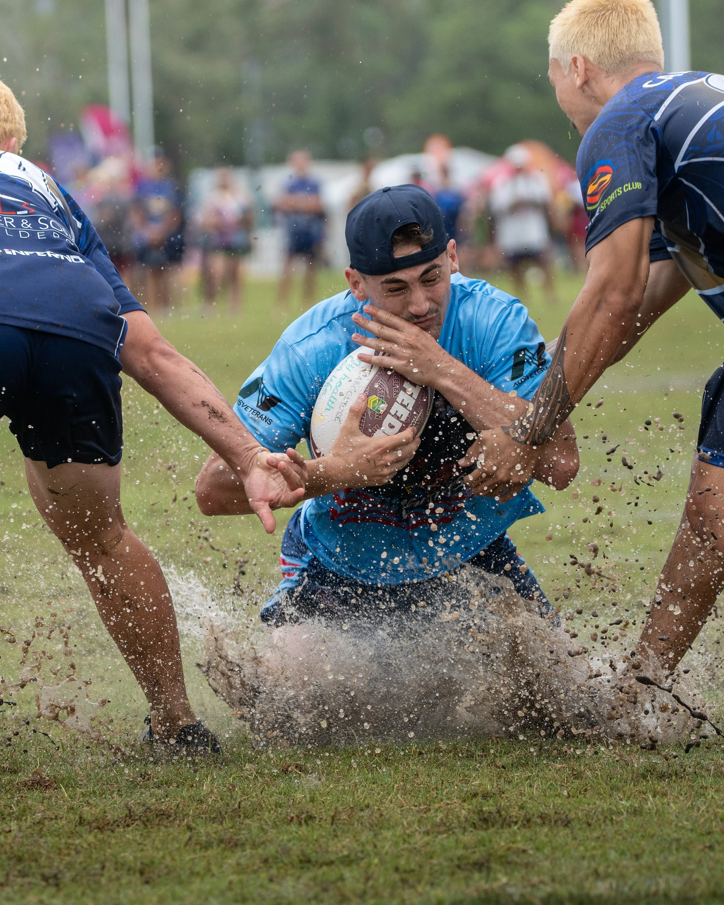 A rugby player in blue uniform with a backwards cap is tackled on a muddy field while holding a rugby ball. Two other players are partially visible, and in the background is a crowd of spectators.