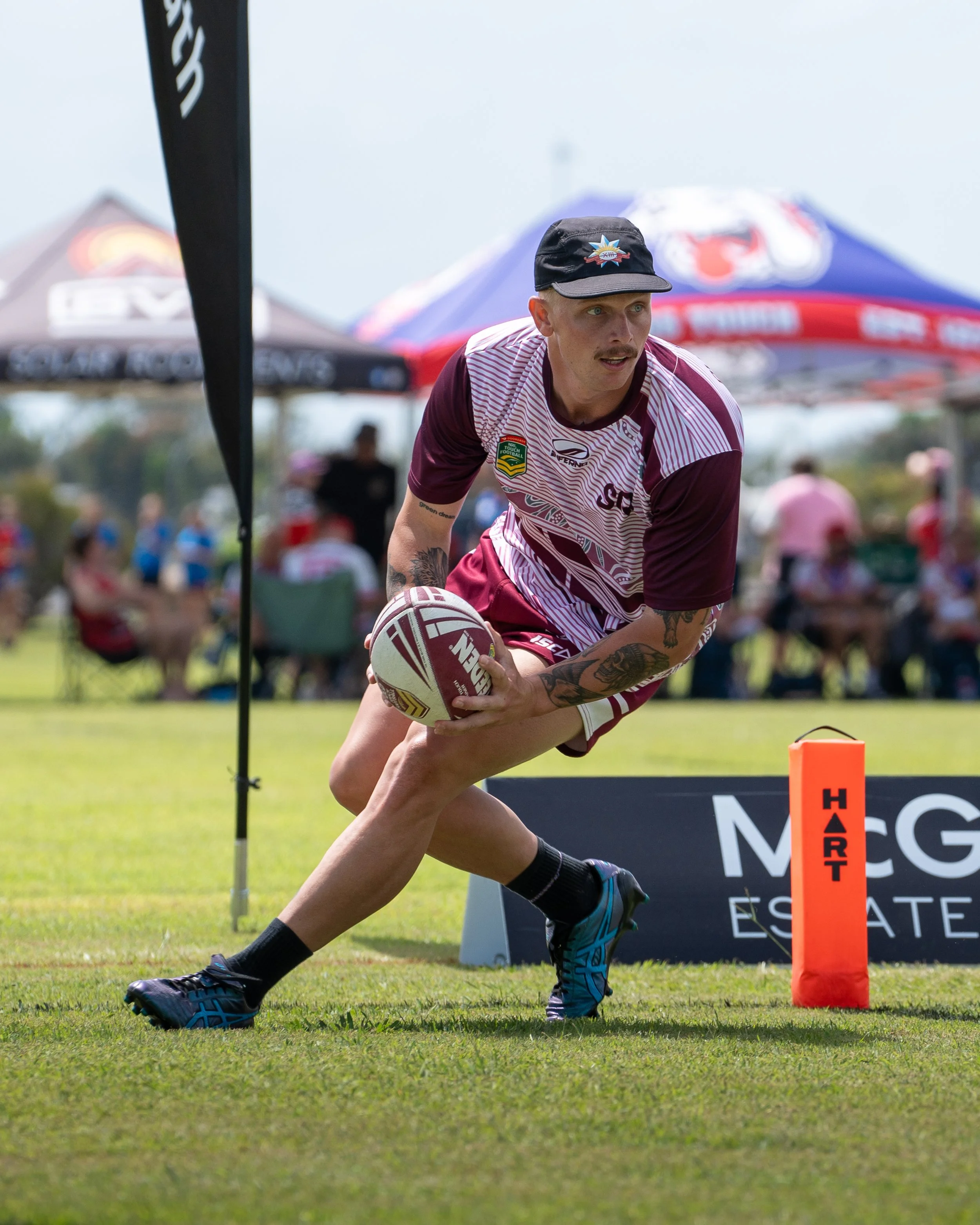 An athlete wearing a maroon and white sports jersey with a patch on the sleeve, black socks, and blue sports shoes is holding a rugby ball and running on a grassy field during a game or practice. There are tents and spectators in the background, and an orange pylon with the word 'HART' is nearby.