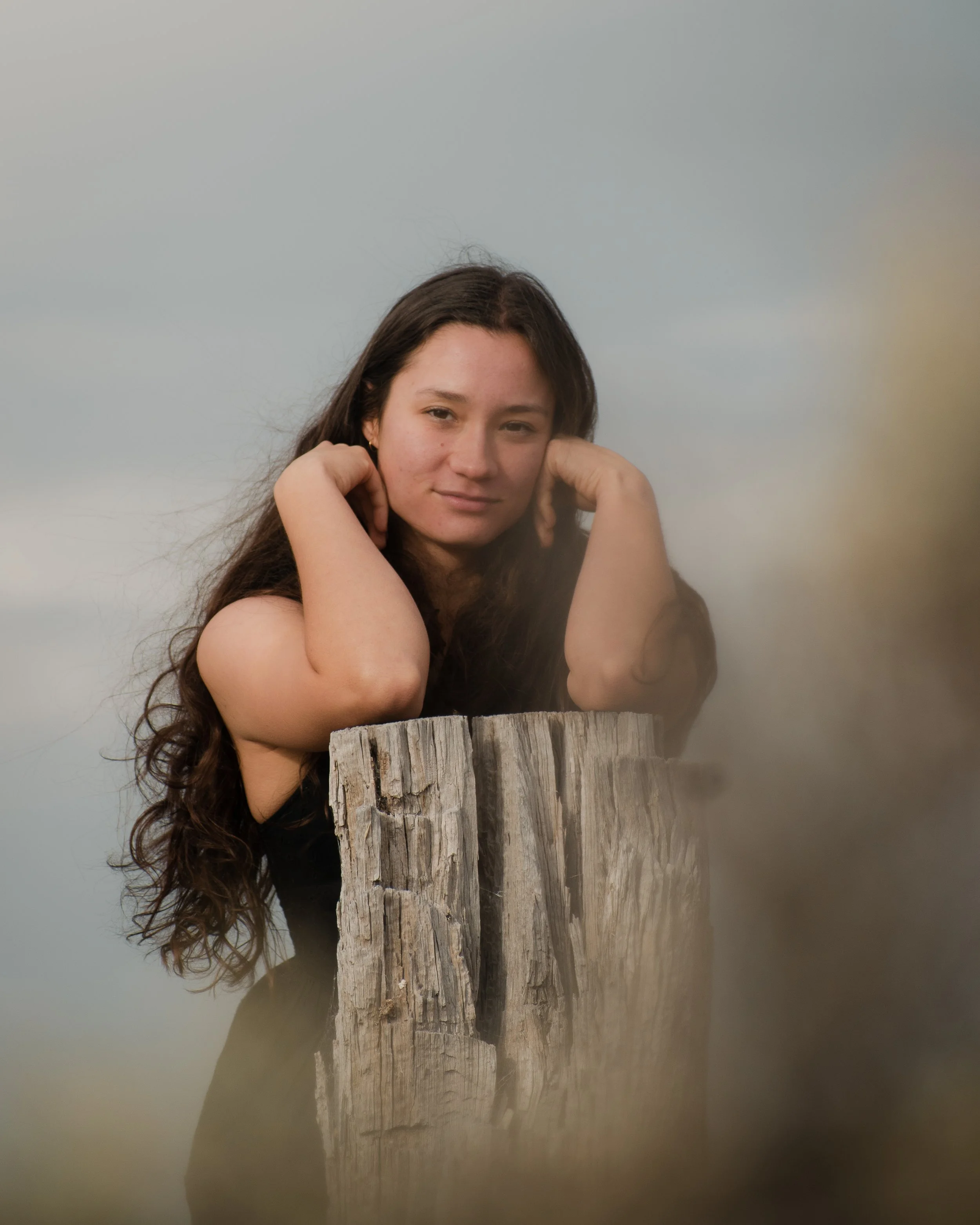 A young woman with long dark hair resting her head on her hands, sitting behind a weathered wooden post, against a plain background.