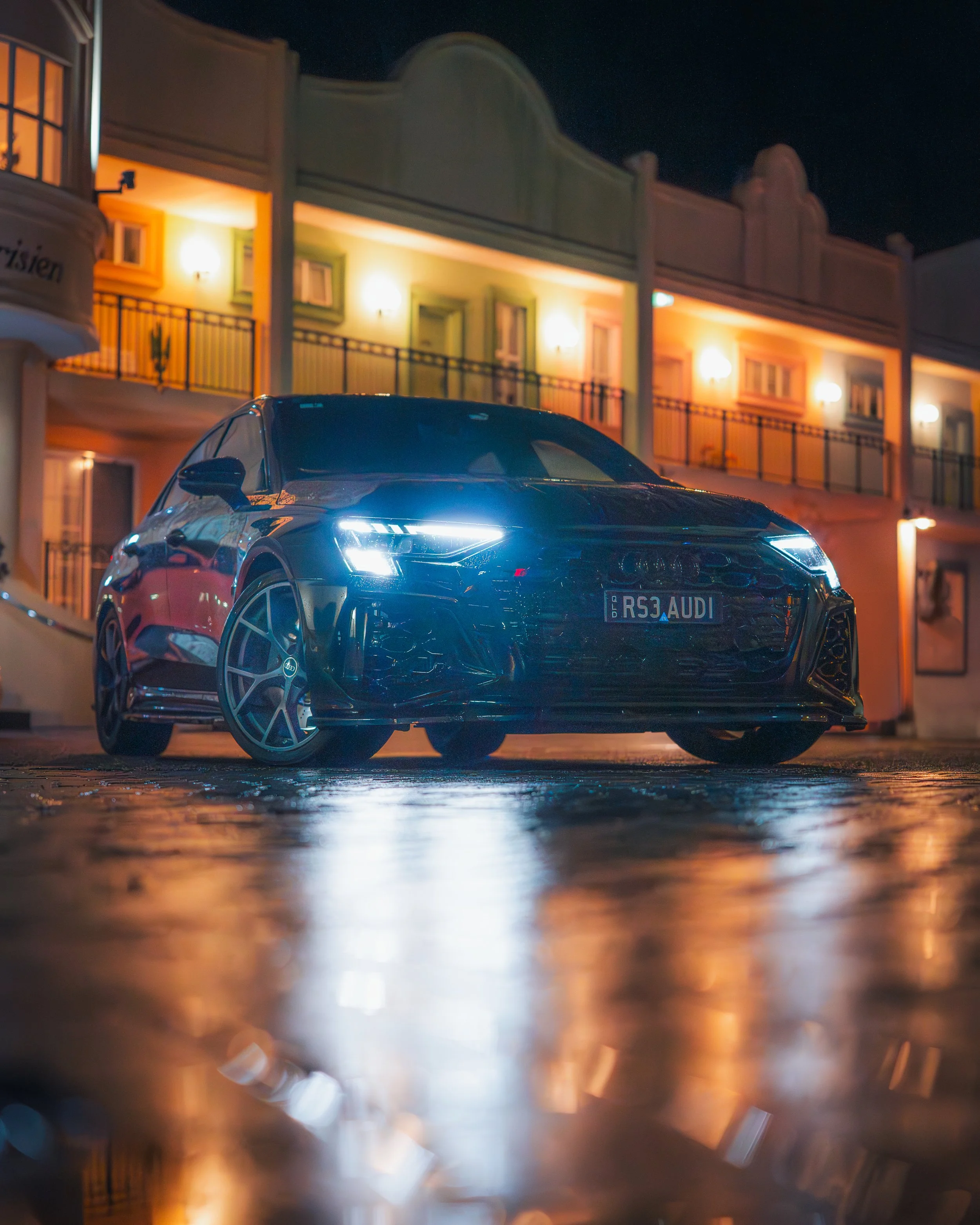 Nighttime photo of a sleek black Audi RS3 car parked on a wet street, reflected on the shiny surface, with colorful buildings illuminated in the background.
