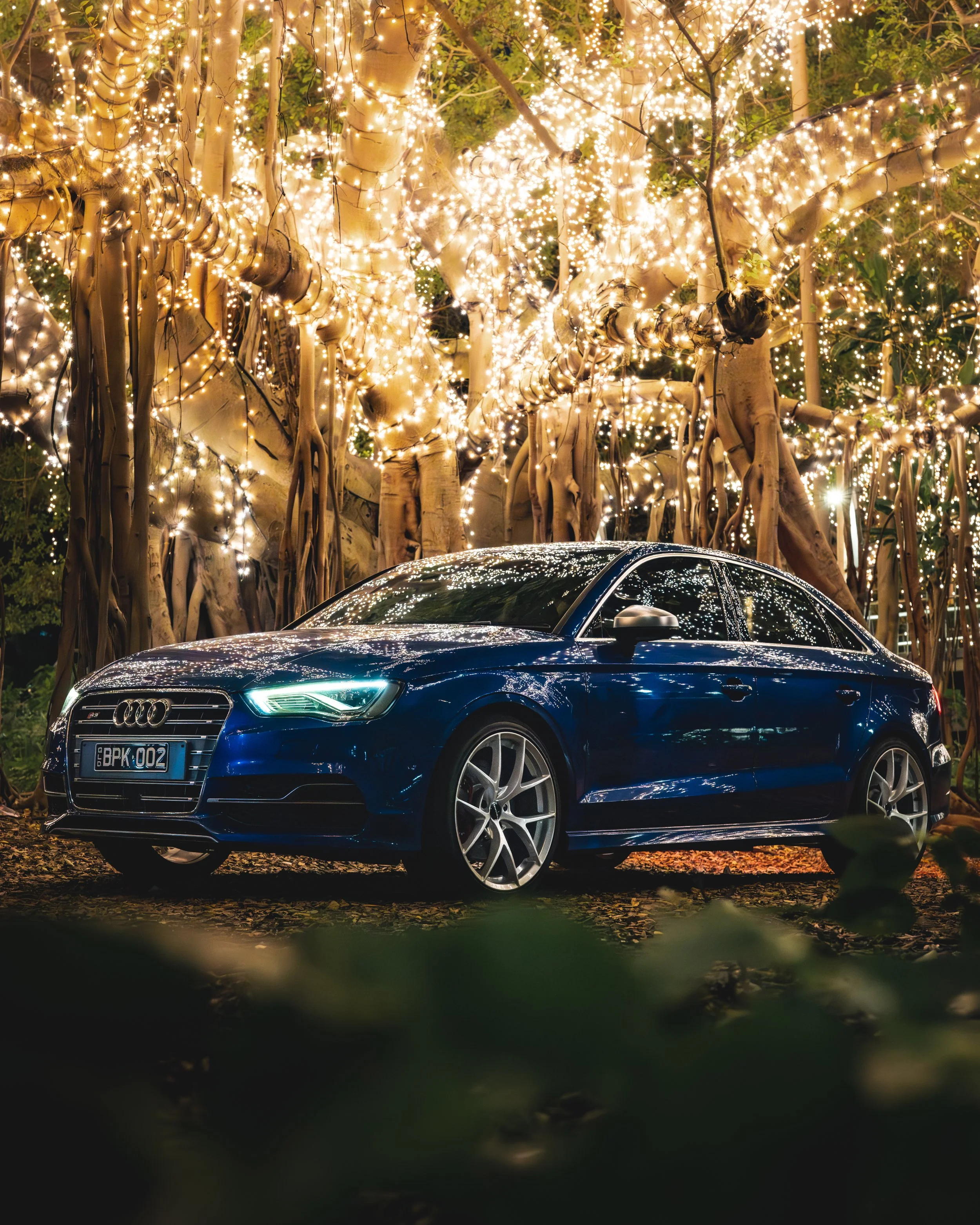 A black Audi sedan parked outdoors at night, with its reflective body and headlights illuminated, surrounded by trees decorated with hanging string lights.