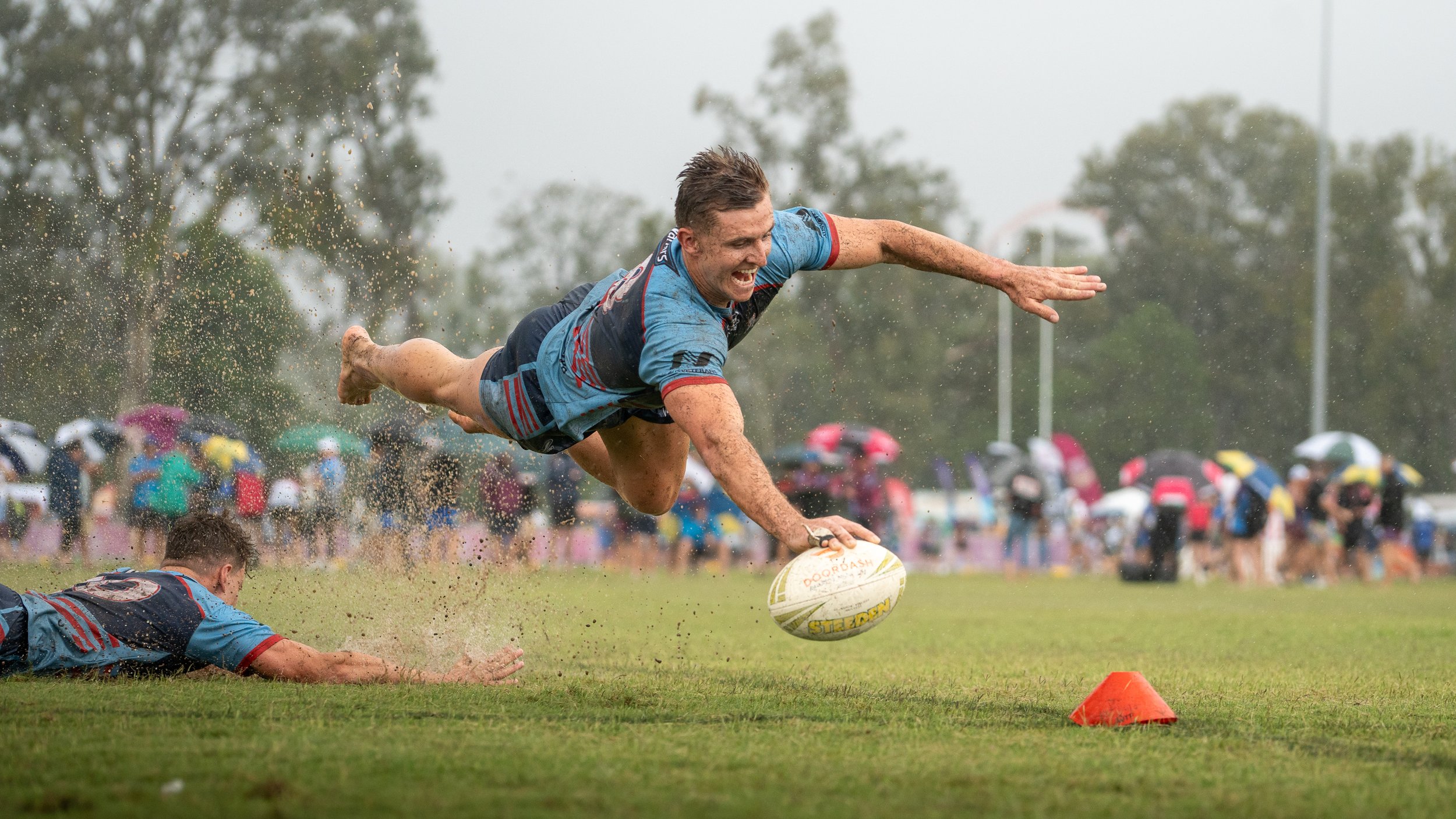 A rugby player diving on a muddy field to catch a rugby ball during a game in the rain with spectators in the background holding umbrellas.