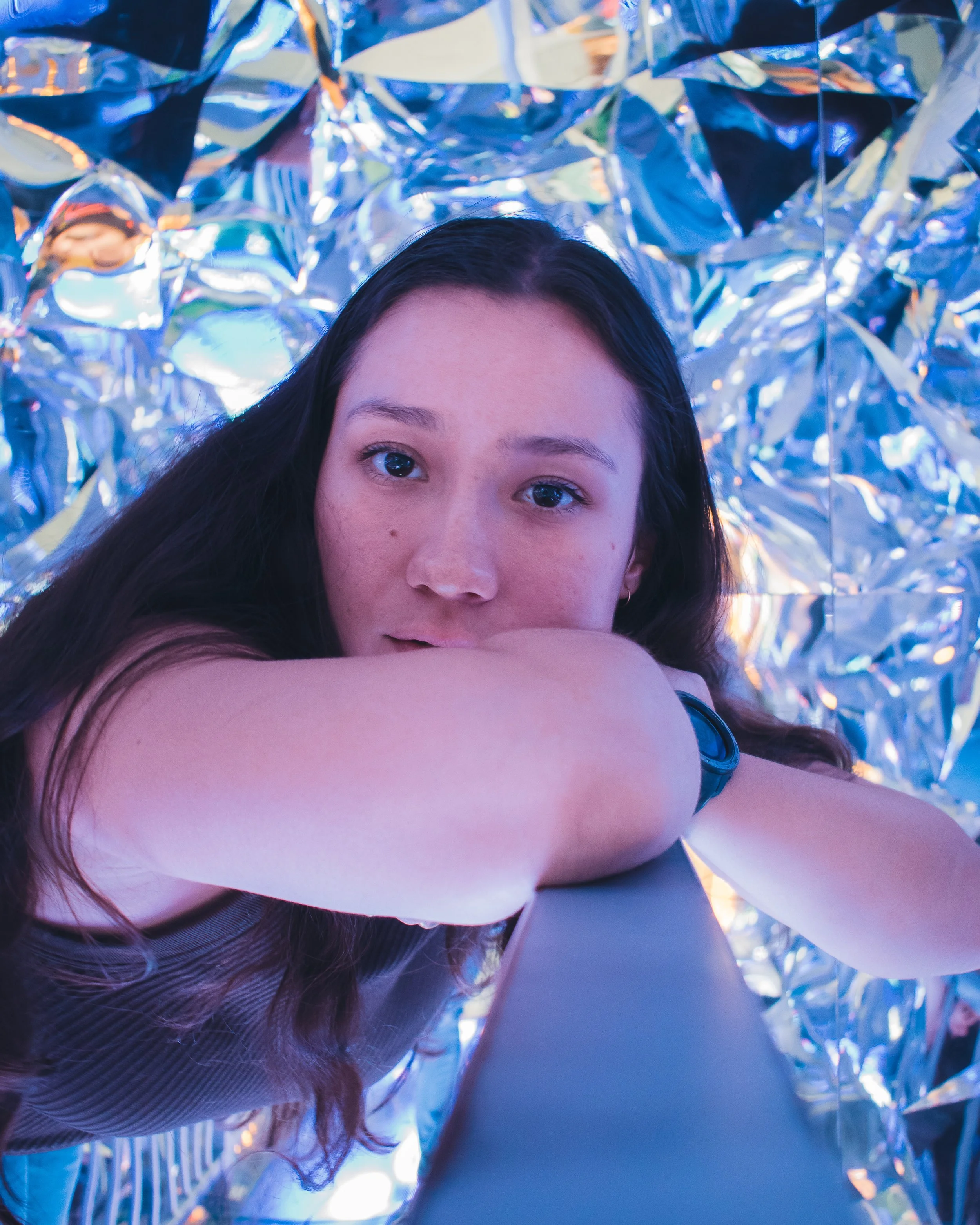A young woman with long dark hair looking directly into the camera, with her arm resting on a flat surface, surrounded by shiny metallic reflective foil decorations.