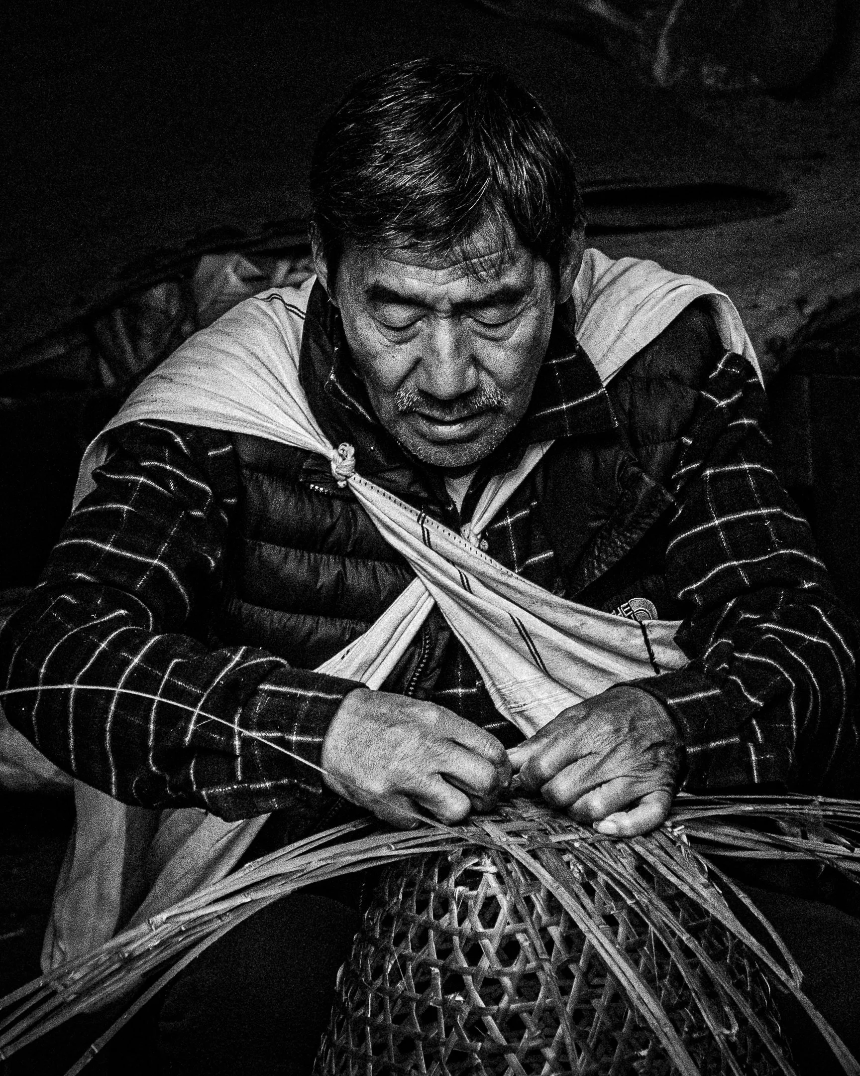 A black and white photograph of a man with short hair and a mustache, wearing a patterned jacket and a light-colored scarf, sitting with his eyes closed and hands clasped together in front of him, surrounded by woven bamboo or wicker structures.