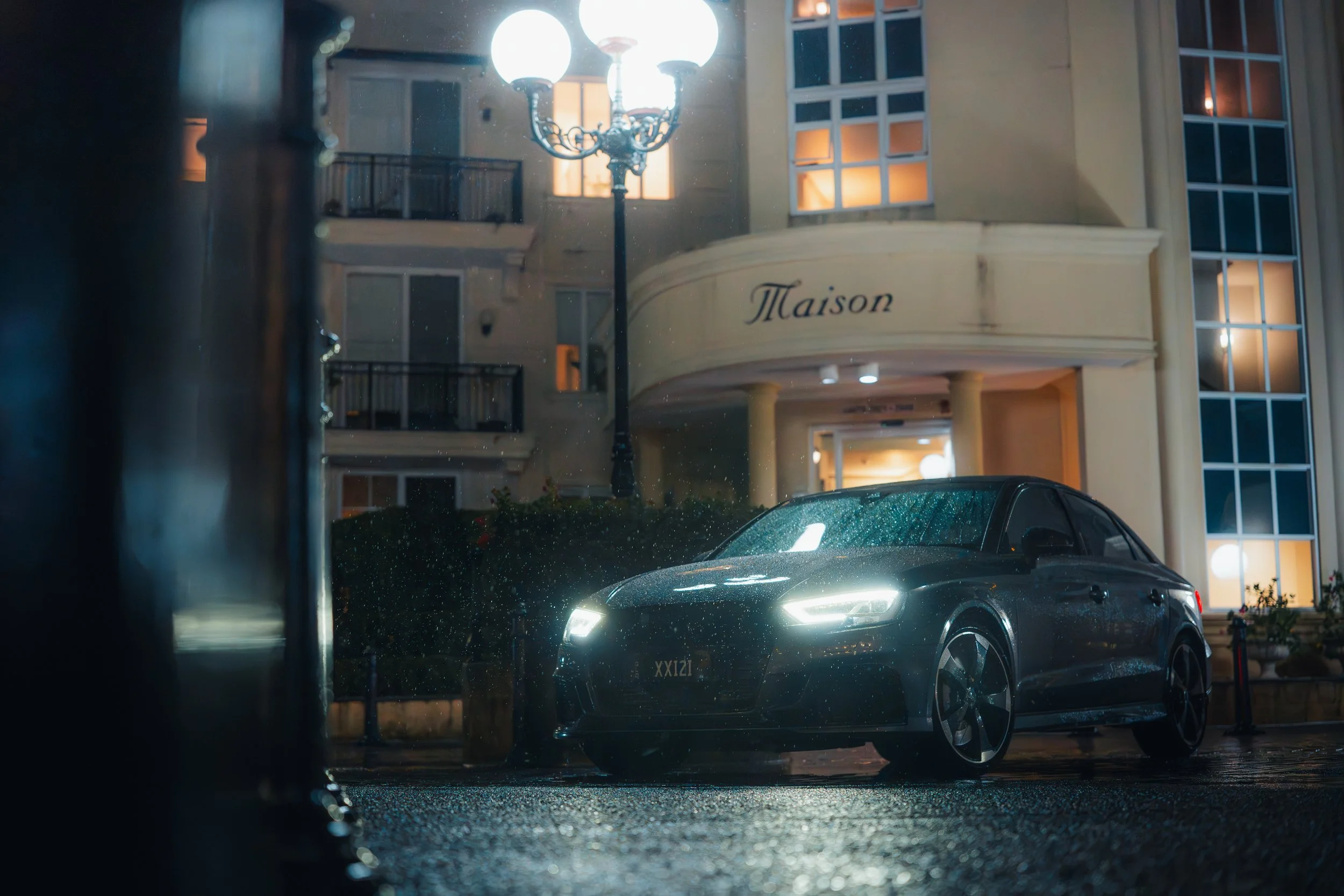 Night scene of a wet street view through a window, with a dark gray car parked on the street, illuminated by a street lamp, and adjacent building with the word 'Maison' on the facade.