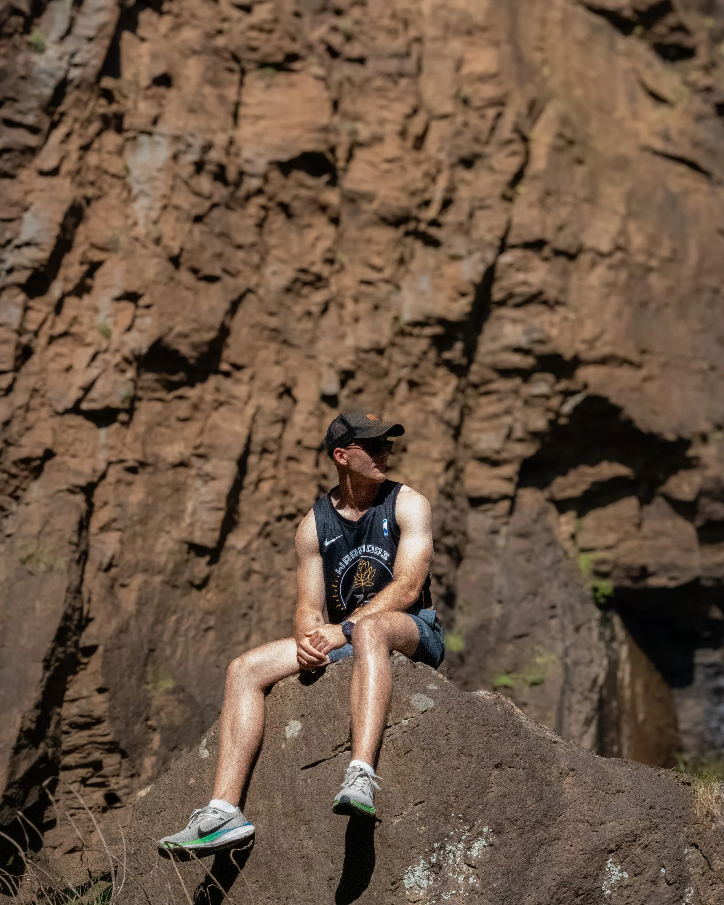 A person sitting on a rock in front of a large rock wall wearing a gray cap, black tank top with Golden State Warriors logo, shorts, and running shoes, with one leg bent and the other extended.