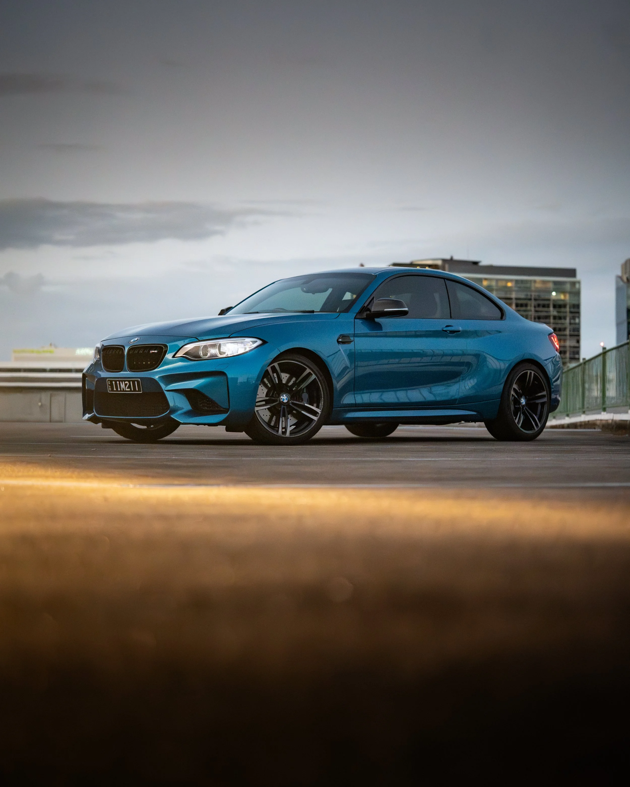 A blue BMW coupe car is parked outdoors in front of a modern glass building with lights on, under a cloudy, overcast sky during dusk or early evening.