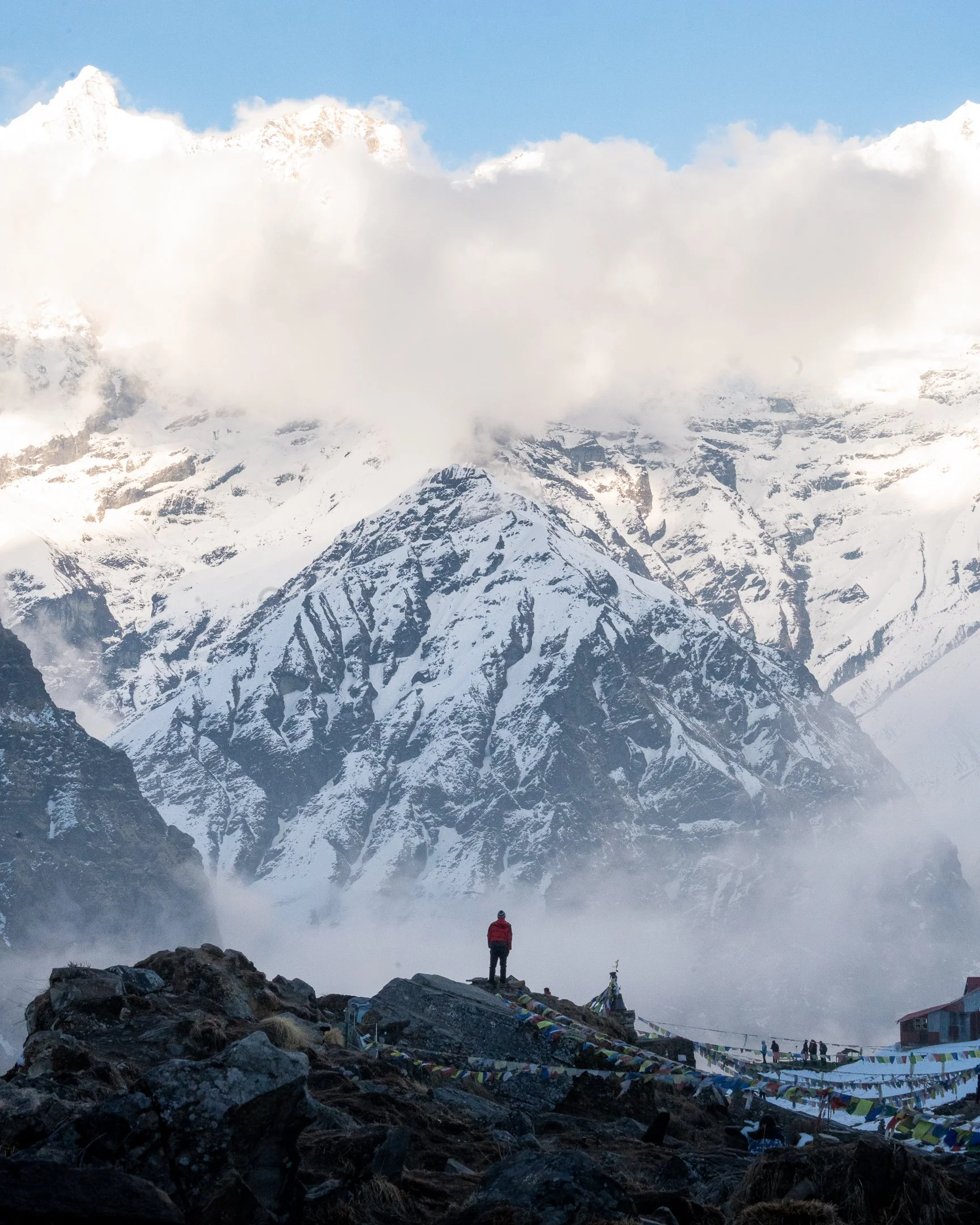 A person standing on rocky ground in front of snow-capped mountains, with prayer flags and clouds covering the peaks.