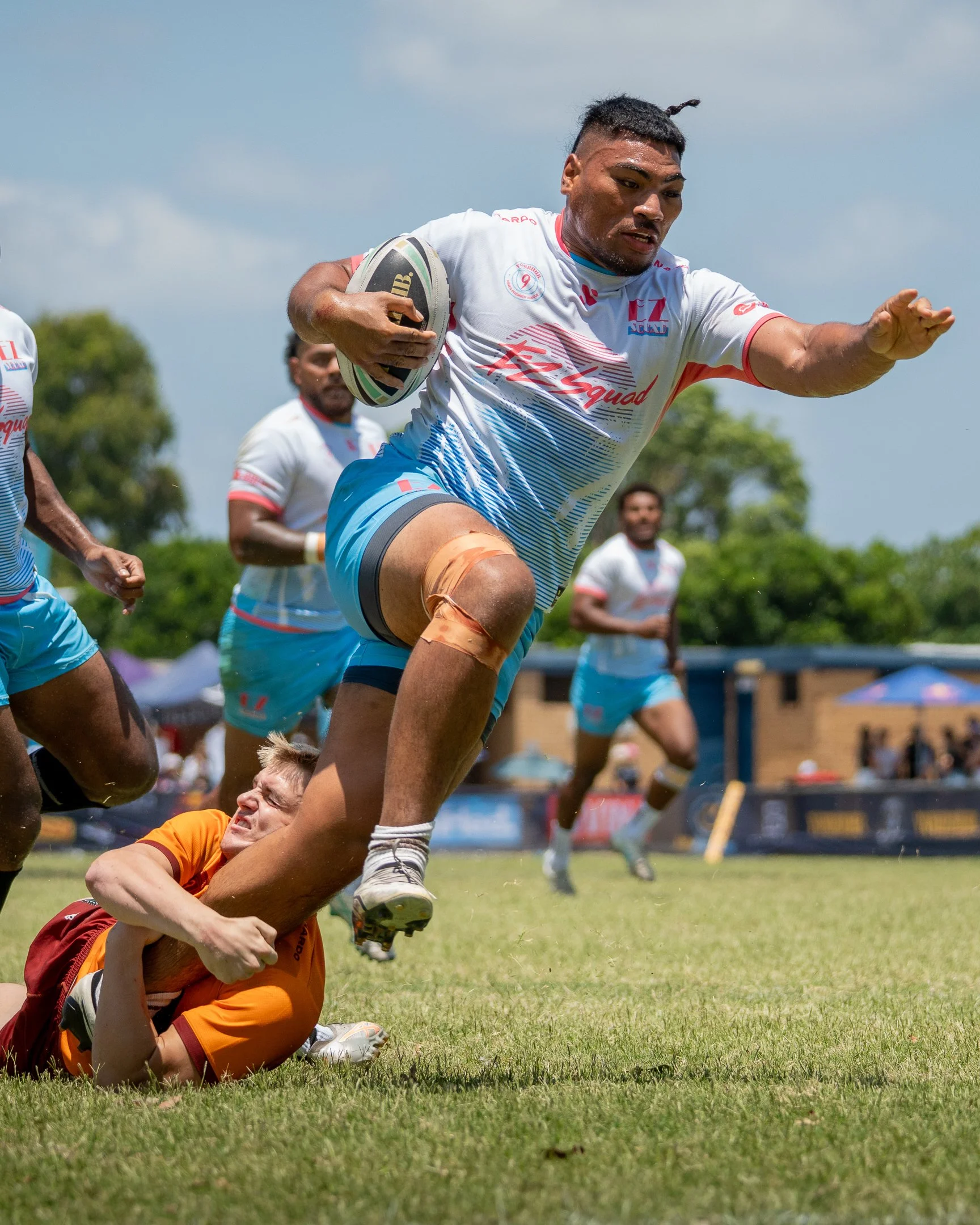 A rugby player in a white jersey running with the ball, evading a tackling opponent in an orange jersey during a game outdoors on a grassy field.