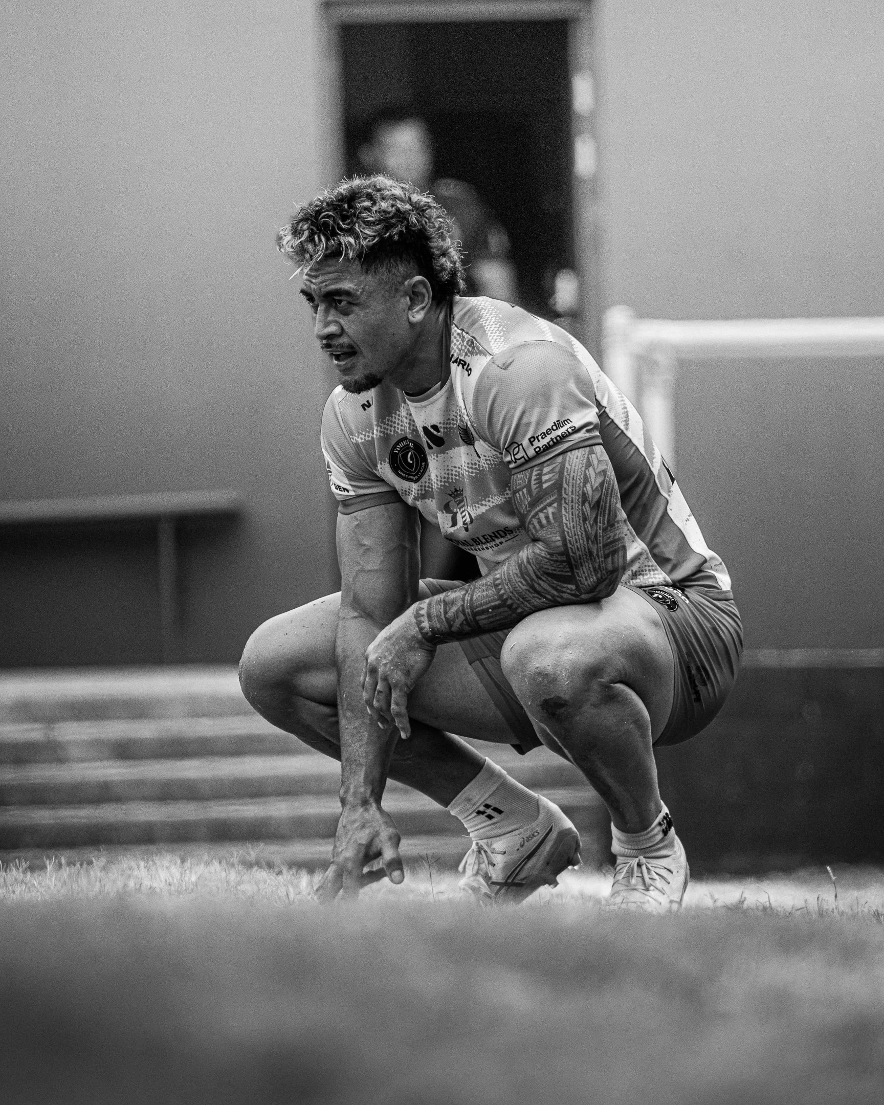 A male athlete crouching on a sports field, wearing a sports jersey, shorts, and sneakers, with focus and effort on his face. The background is blurred.