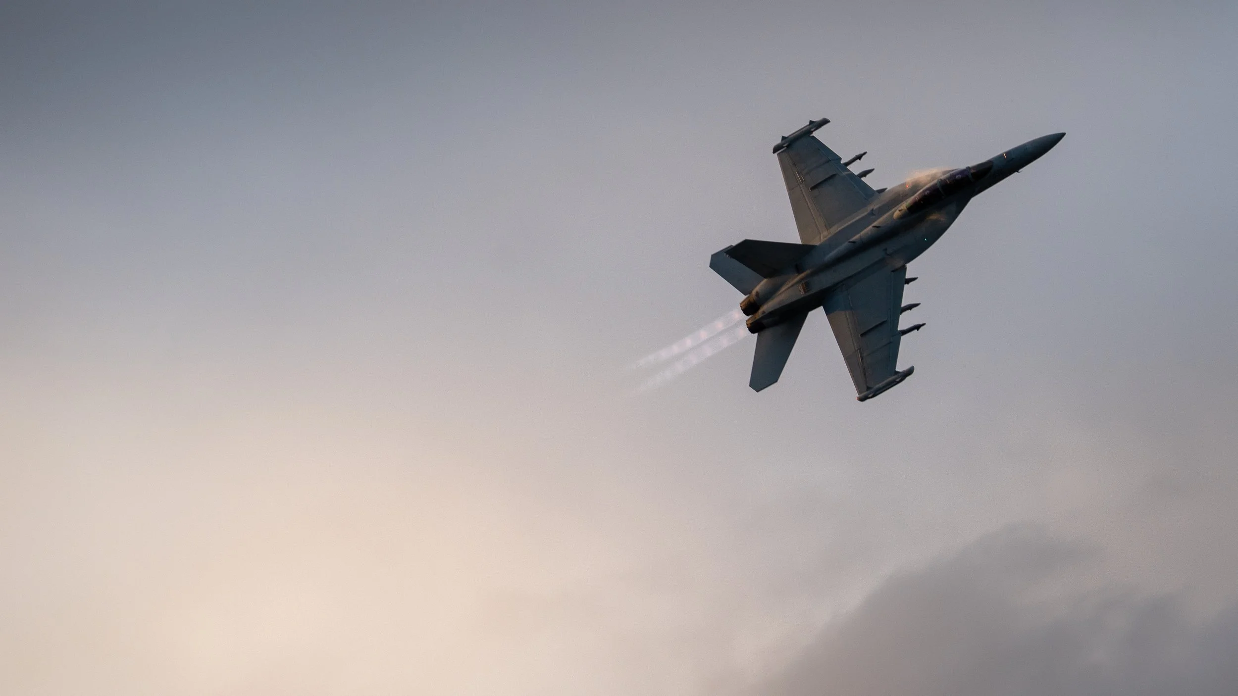 Military fighter jet flying through the sky at an angle, with vapor trails behind it, during dusk or dawn.