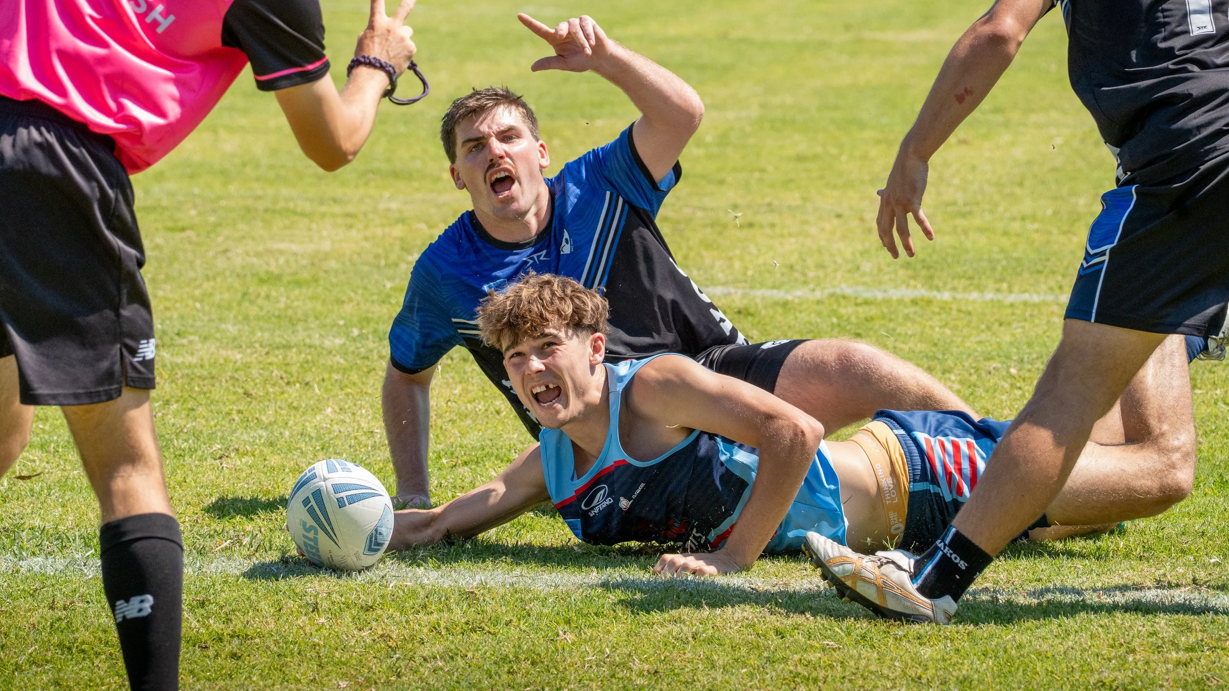 Rugby players in action during a game on a grassy field, with one player reaching for the ball while others are on the ground or standing around him.