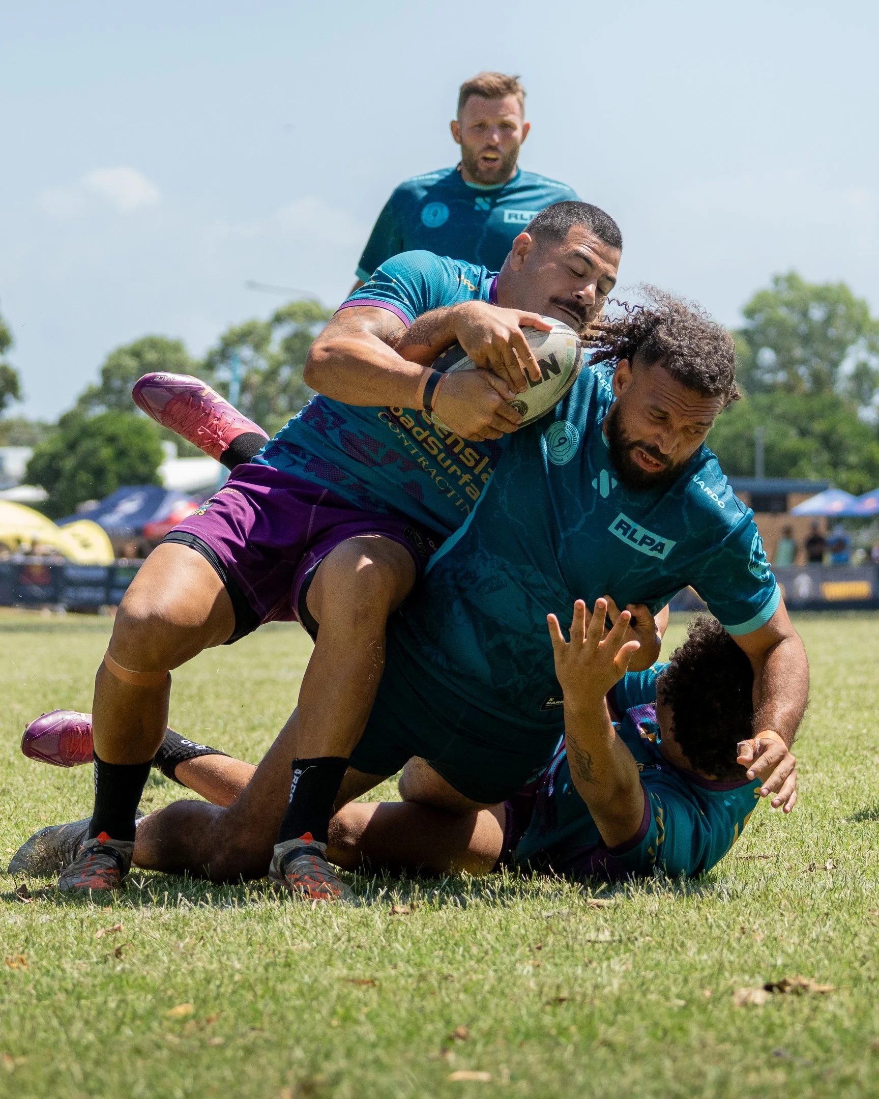 Two rugby players are engaging in a tackle during a match, with one player on top of the other holding a rugby ball, while a third player observes in the background.