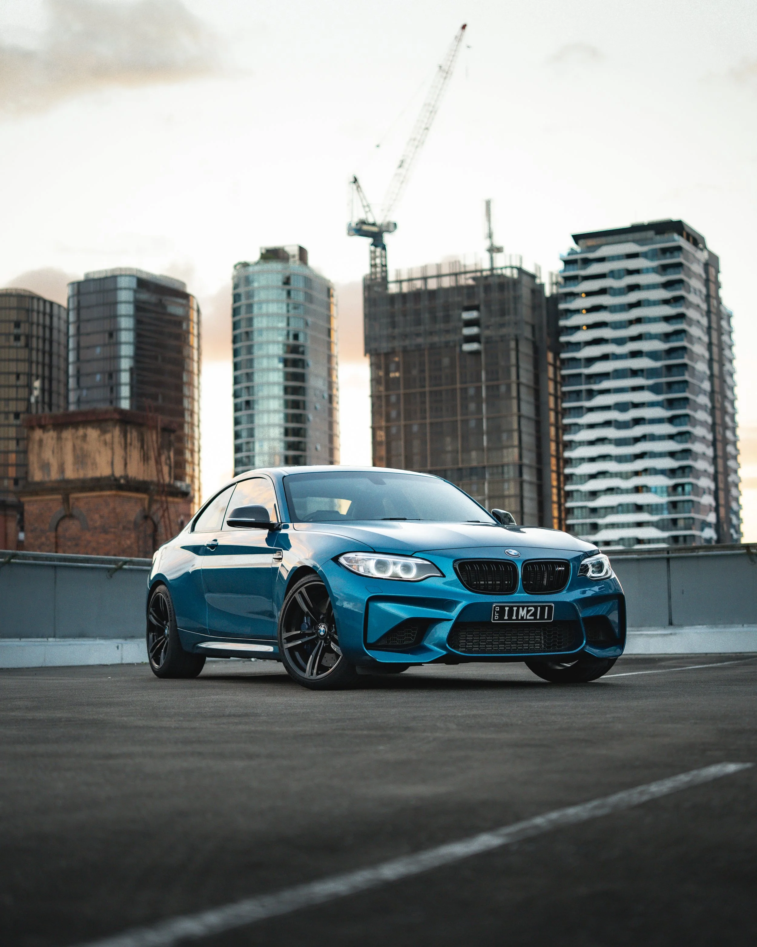 A blue BMW sports car parked on a rooftop parking lot with high-rise buildings and a construction crane in the background during sunset or early evening.