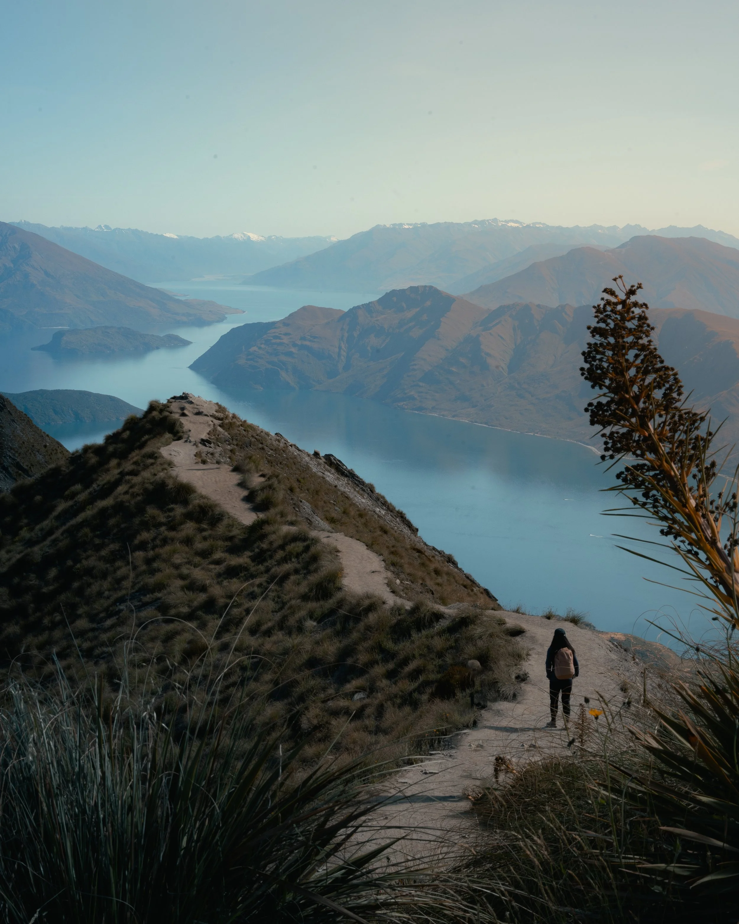 Hiker walking on a dirt path through hills and mountains near a large lake, with snow-capped peaks in the distance.