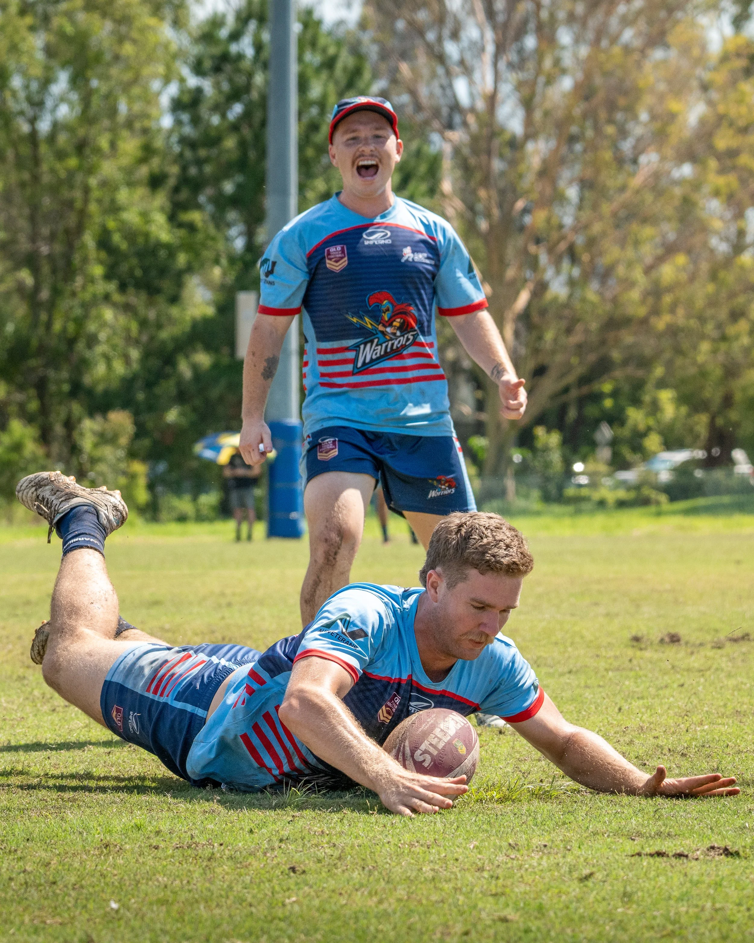 Two male rugby players in blue and red uniforms are playing on a grassy field; one is on the ground holding a rugby ball, while the other is standing and celebrating.