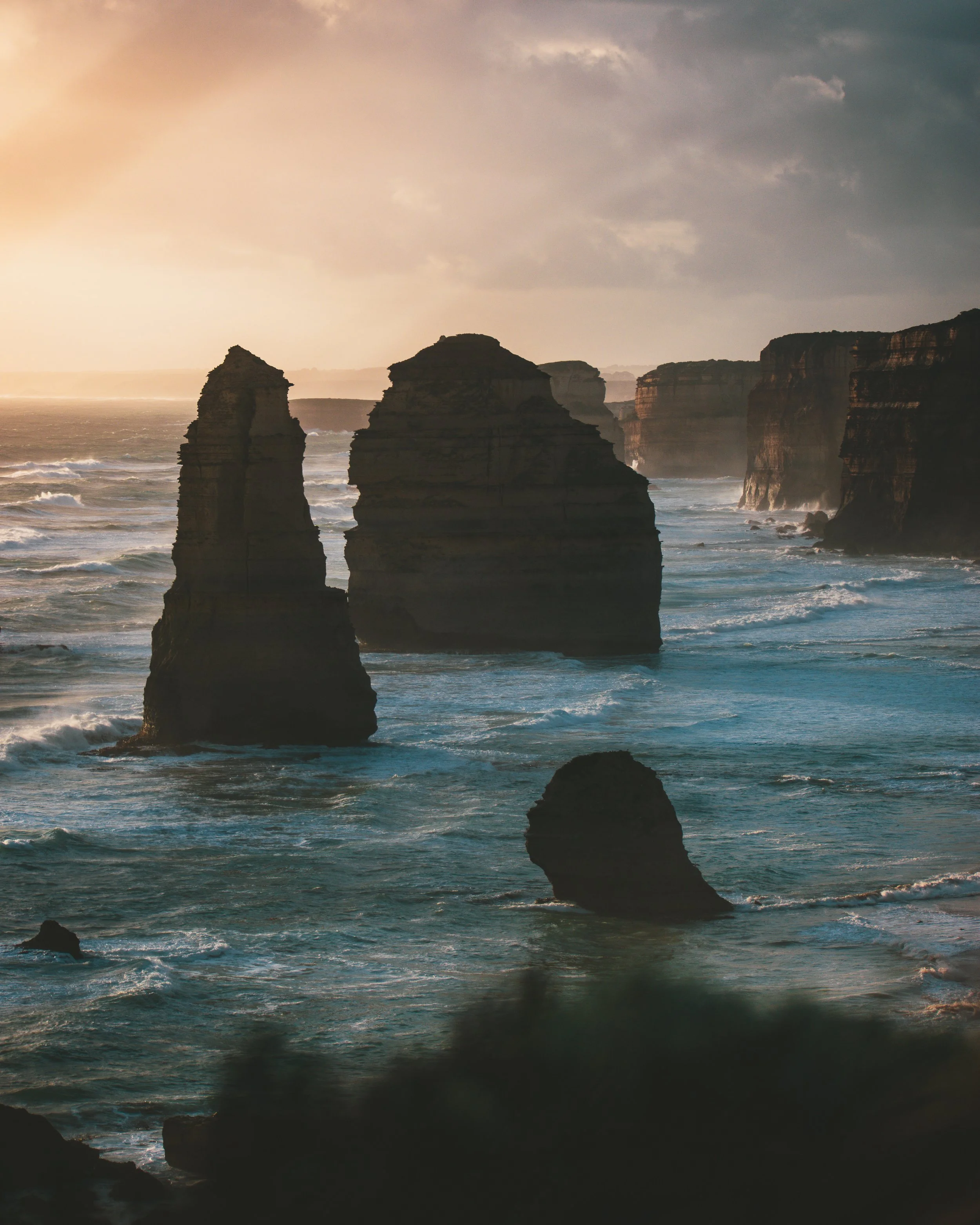 Ocean waves crashing against dark, rugged rock formations along a coastline during sunset, with a cloudy sky overhead.