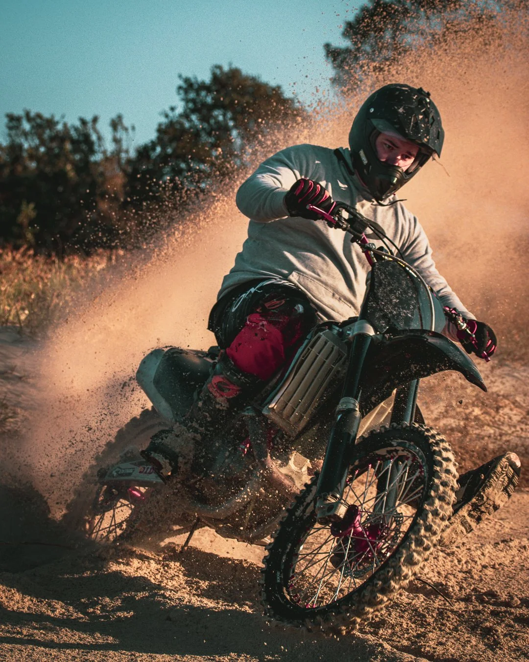 Person riding a dirt bike on sandy terrain with dust and spray flying, wearing a helmet and protective gear.