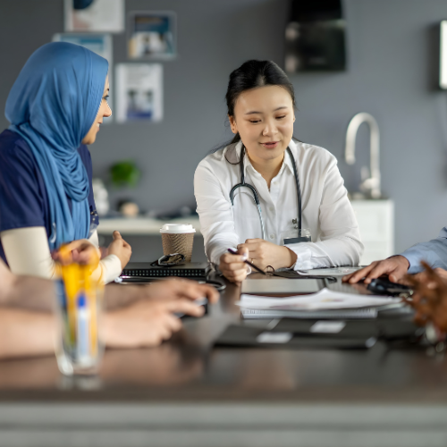 A female doctor in medical attire discussing with a woman wearing a hijab and others in a medical office conference room.