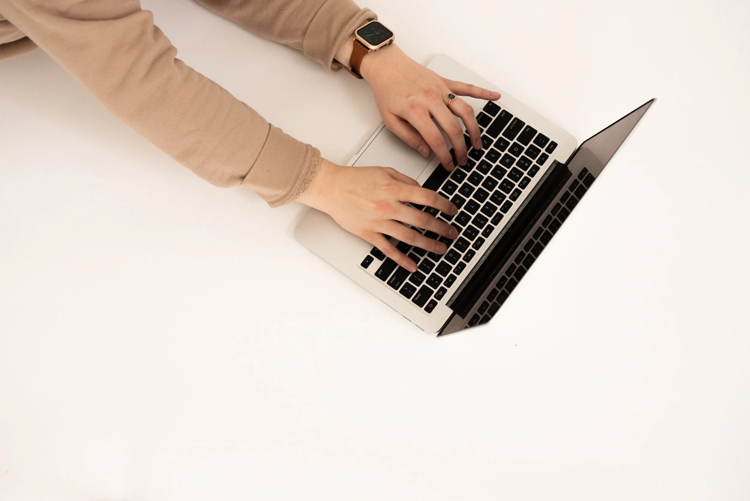A person wearing a beige long-sleeve shirt and a smartwatch is typing on a silver laptop with black keys on a white surface.