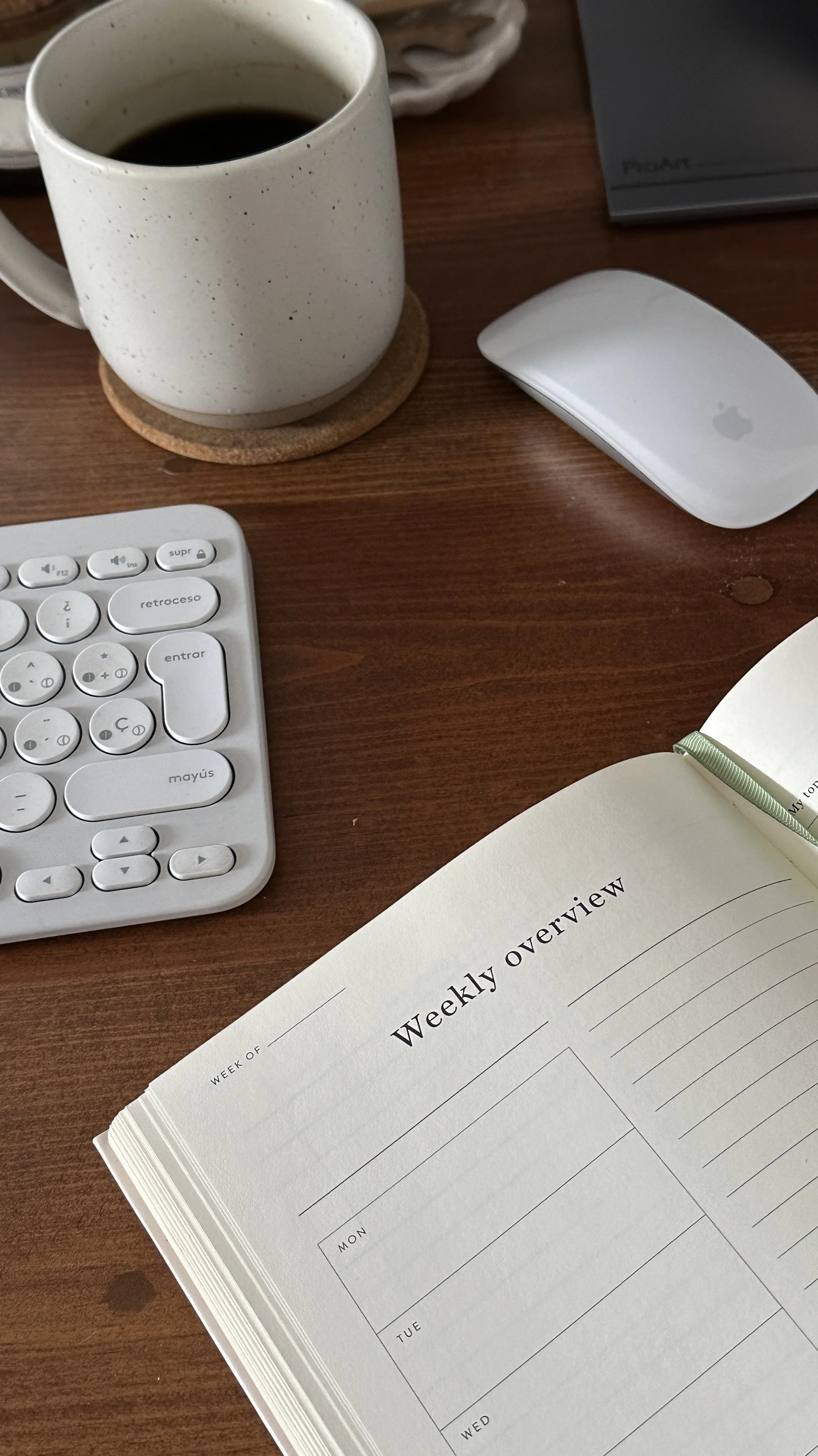 A workspace featuring a white mug of black coffee on a cork coaster, an Apple Magic Mouse, a partially open weekly planner, and a keyboard on a wooden desk.
