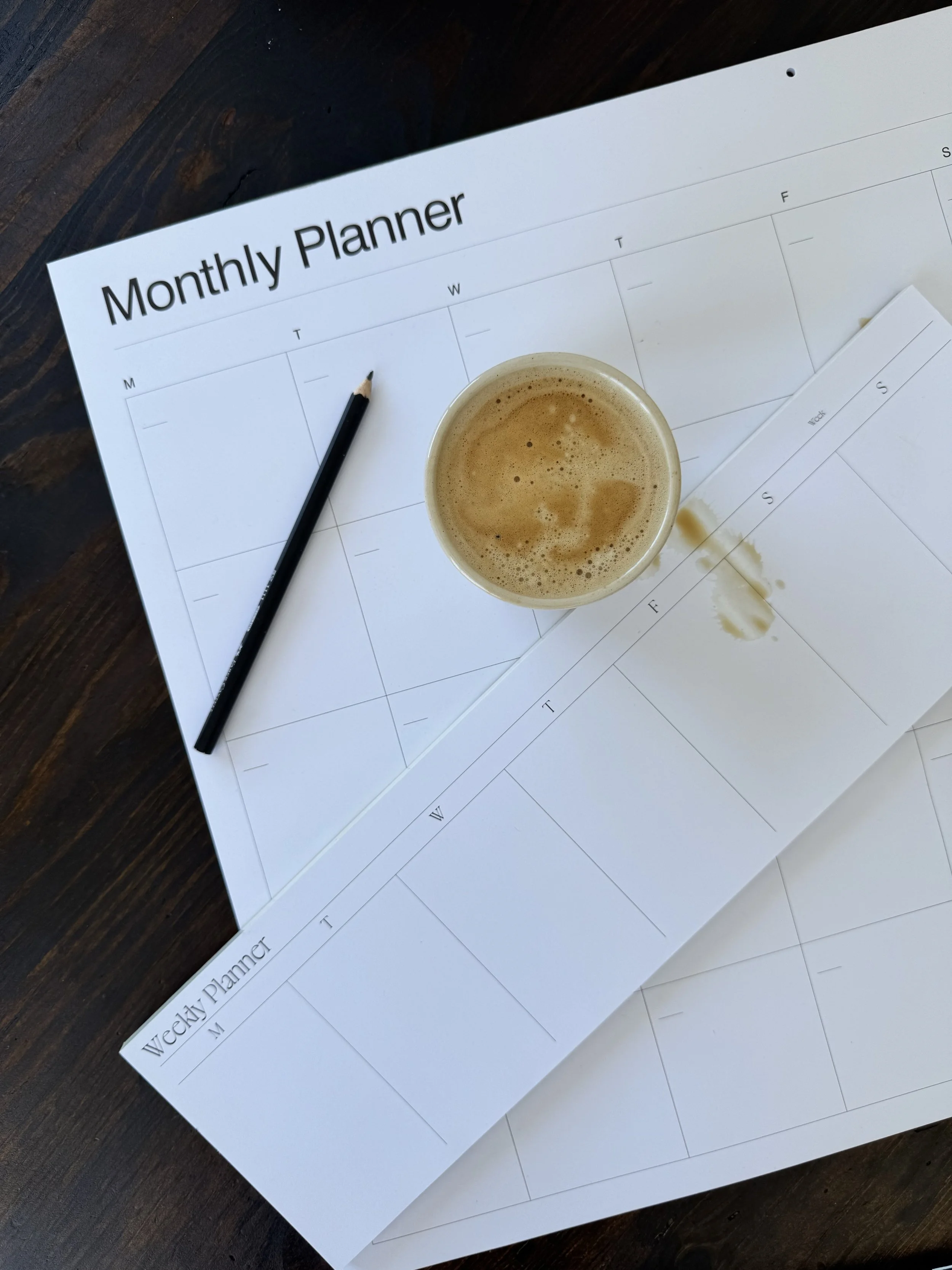 A weekly planner paper labeled 'Monthly Planner' on top of a paper calendar with coffee on it, placed on a wooden surface.