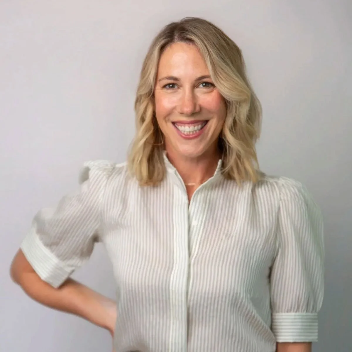 Woman with blonde hair smiling, wearing a white pinstripe blouse, standing against a plain light-colored background.