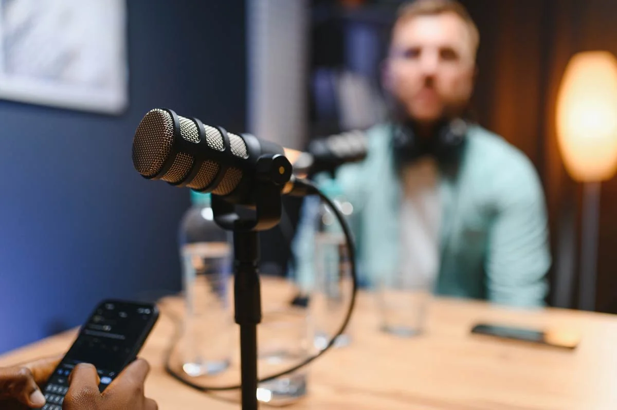 Close-up of a microphone on a stand in front of a person sitting at a table, with a blurred person in the background.