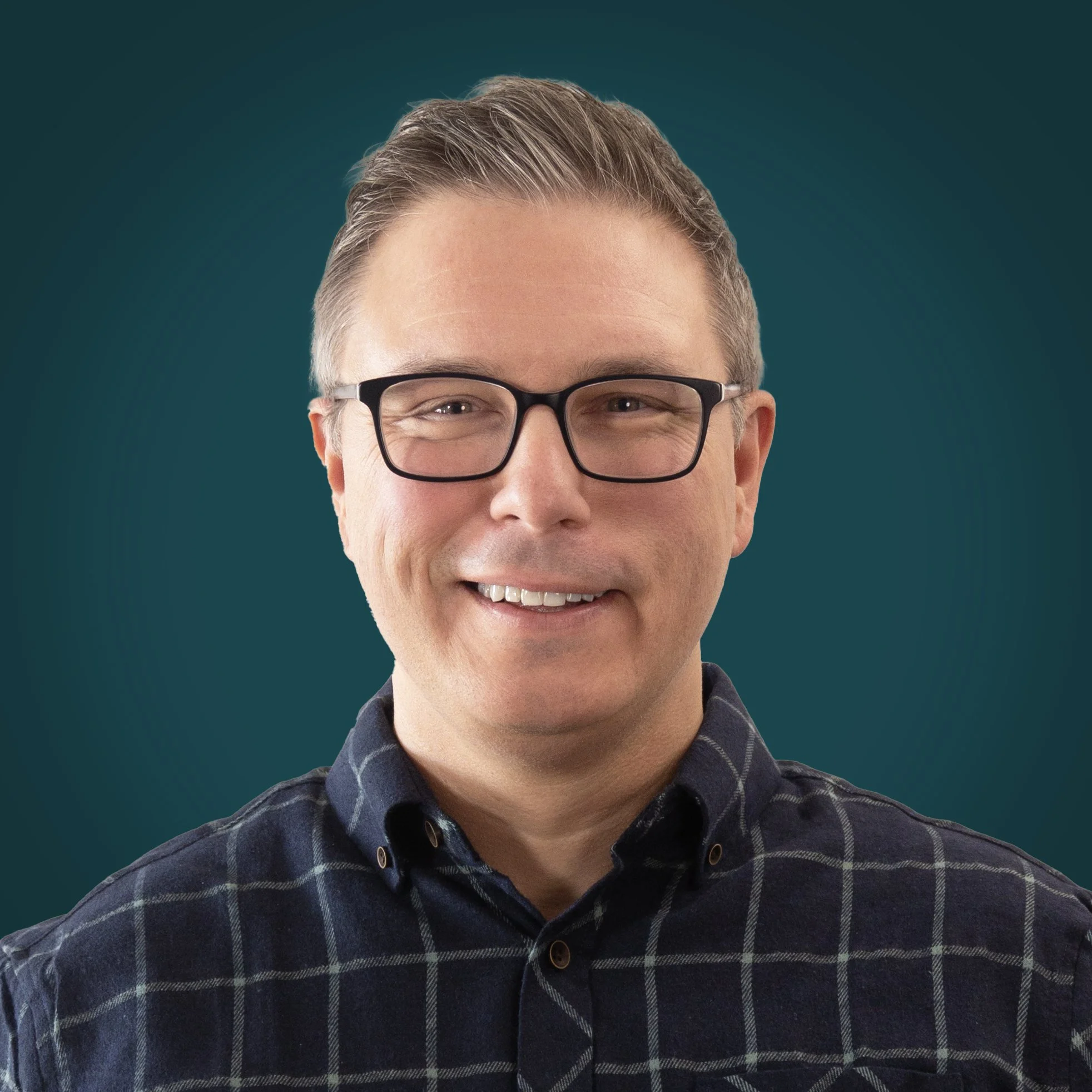Close-up of a smiling man wearing glasses with a teal background and a dark plaid shirt.