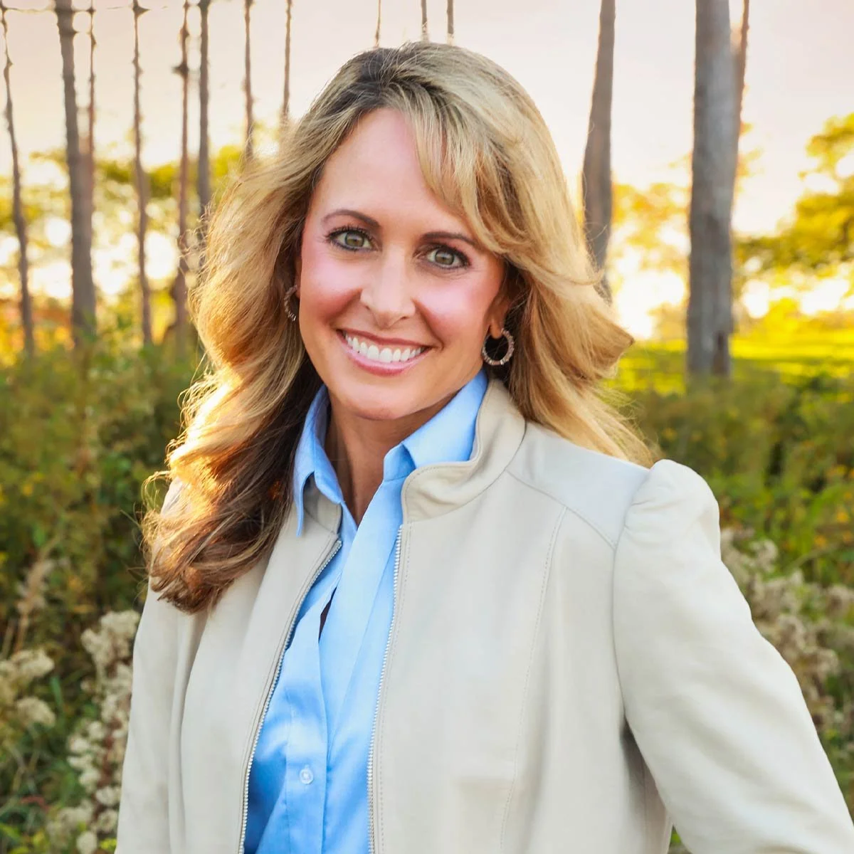 A woman with blonde hair, blue eyes, and a bright smile standing outdoors in a forest during fall. She is wearing a light-colored jacket over a blue shirt, with earrings, and has a background of trees and greenery.