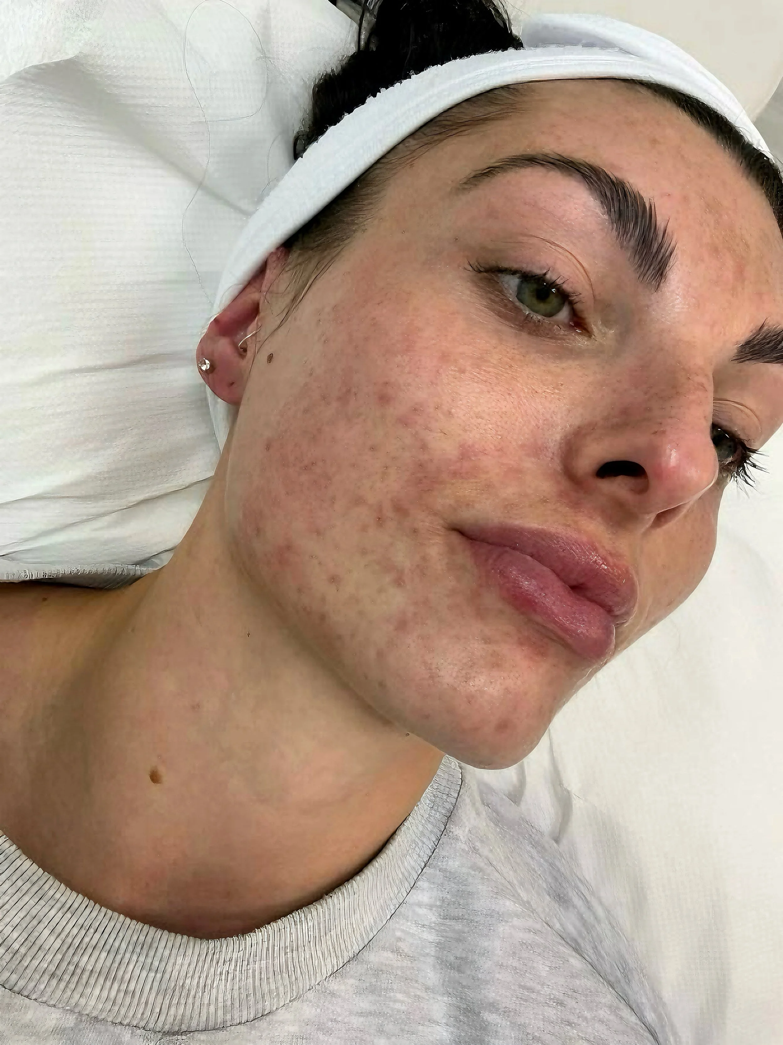 Close-up of a woman lying down, with her face turned slightly to the left, showing her eye, eyebrow, and cheek, wearing makeup, and her dark hair pulled back with a white headband.