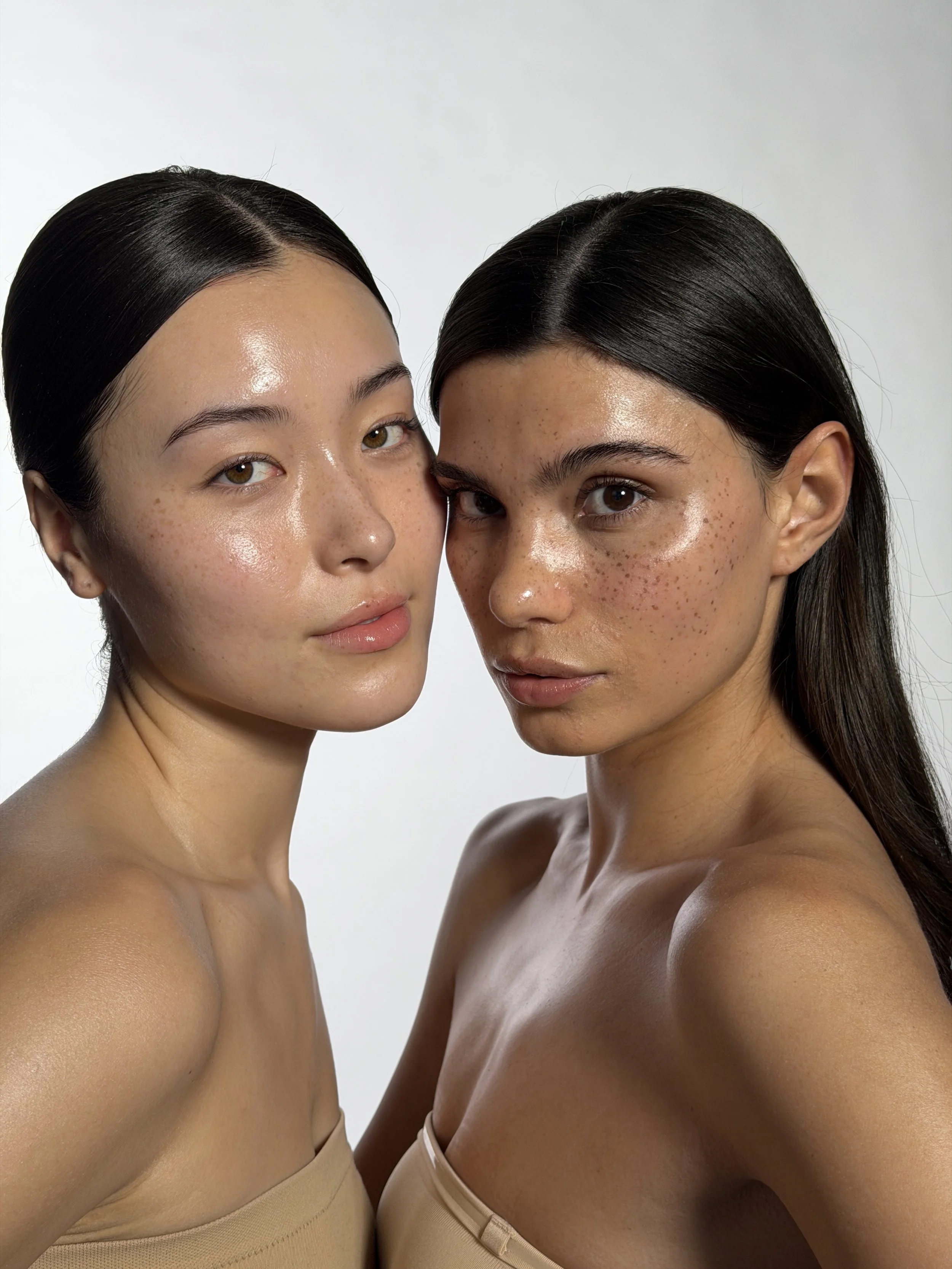 Close-up photograph of two women with dark hair, one with light brown skin and the other with darker skin, both wearing beige strapless tops, standing face-to-face against a white background.