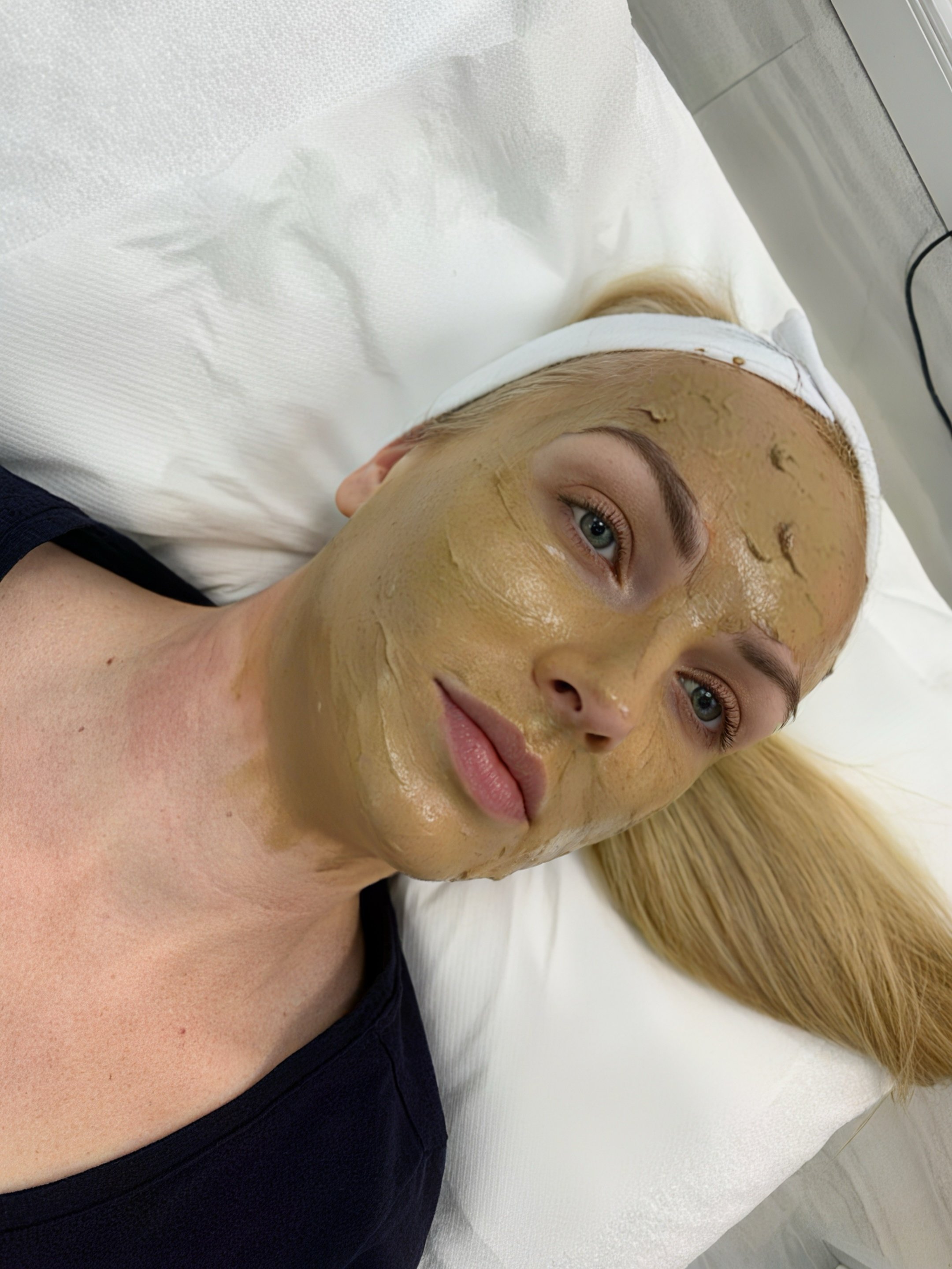 A woman with blonde hair and light-colored eyes lying on a bed, receiving a facial treatment with a greenish-brown facial mask. She is wearing a headband and a dark shirt.