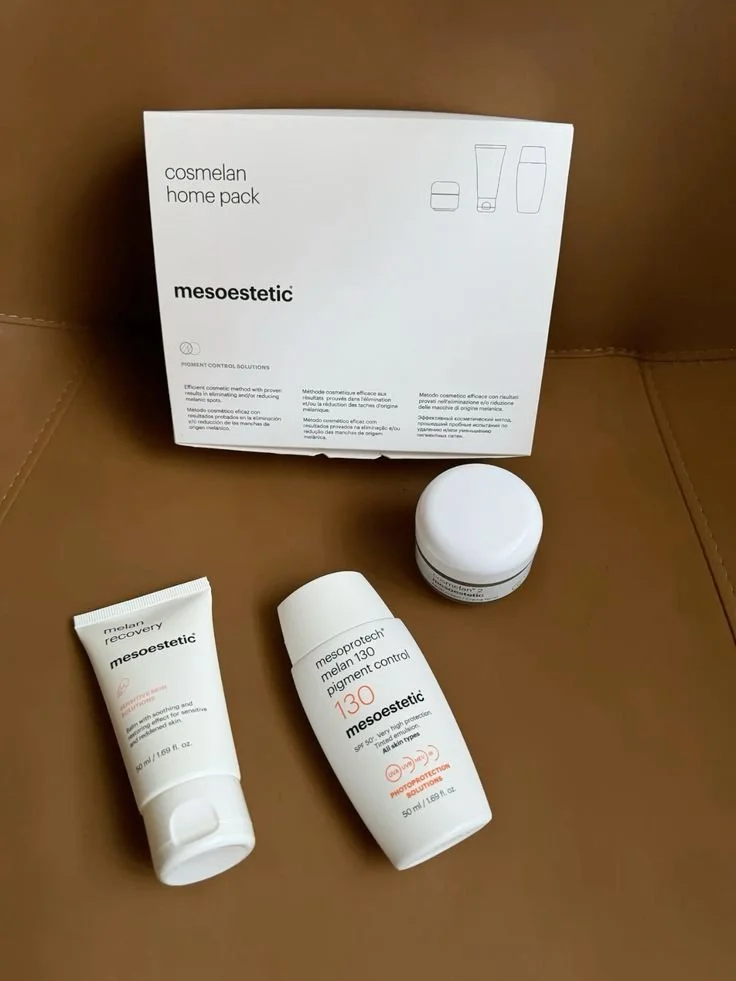 A home skincare kit including a box labeled 'cosmelan home pack,' a small tube labeled 'mesoestetic melas recovery,' a white bottle labeled 'mesoprotech melanin 130 pigment control,' and a small white container, all placed on a brown surface.