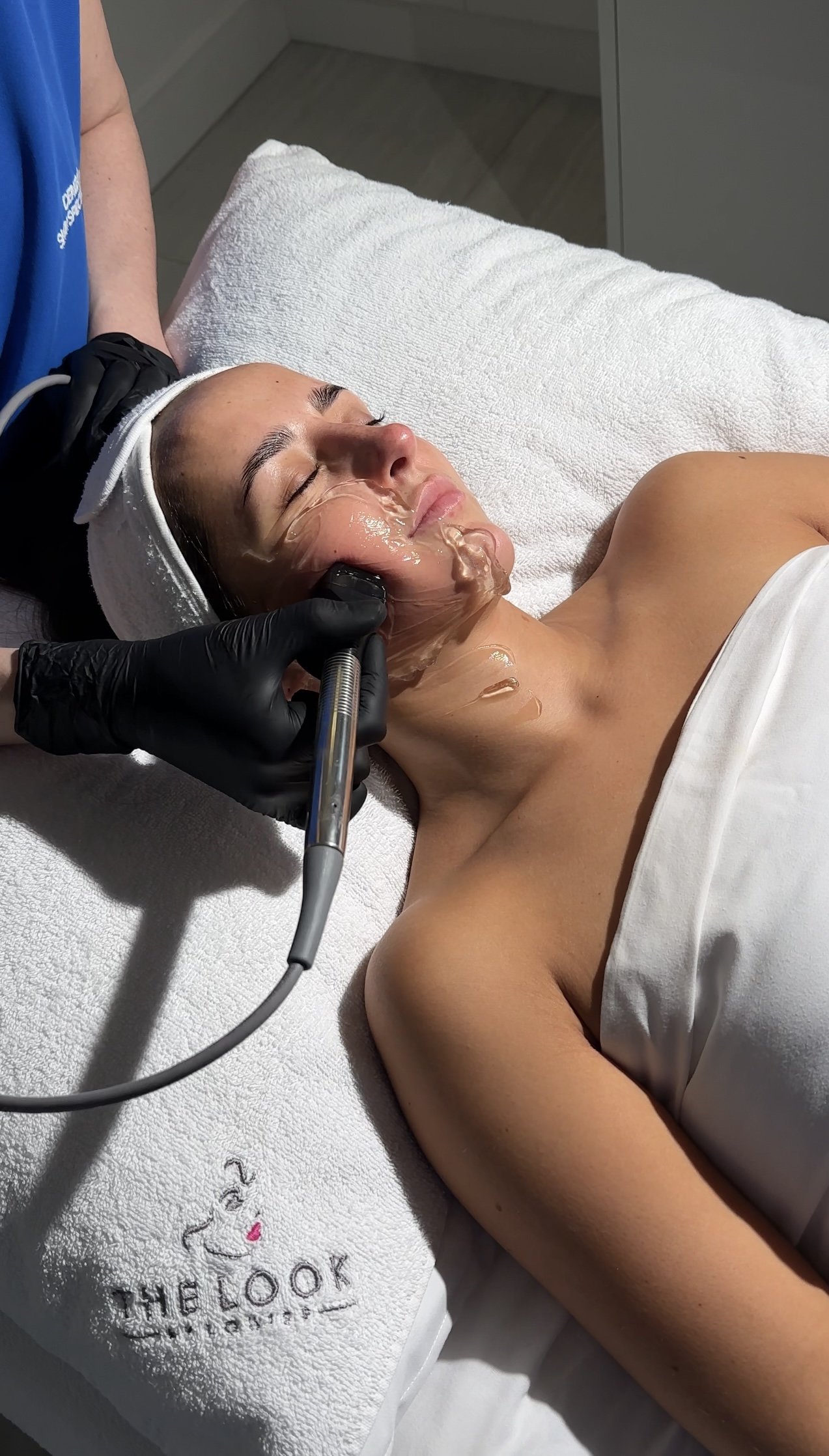A woman receiving a facial treatment at a spa, lying on a treatment bed with a towel embroidered with 'THE LOOK' logo. She has her eyes closed and a mask or facial product is being applied to her face with a handheld device.