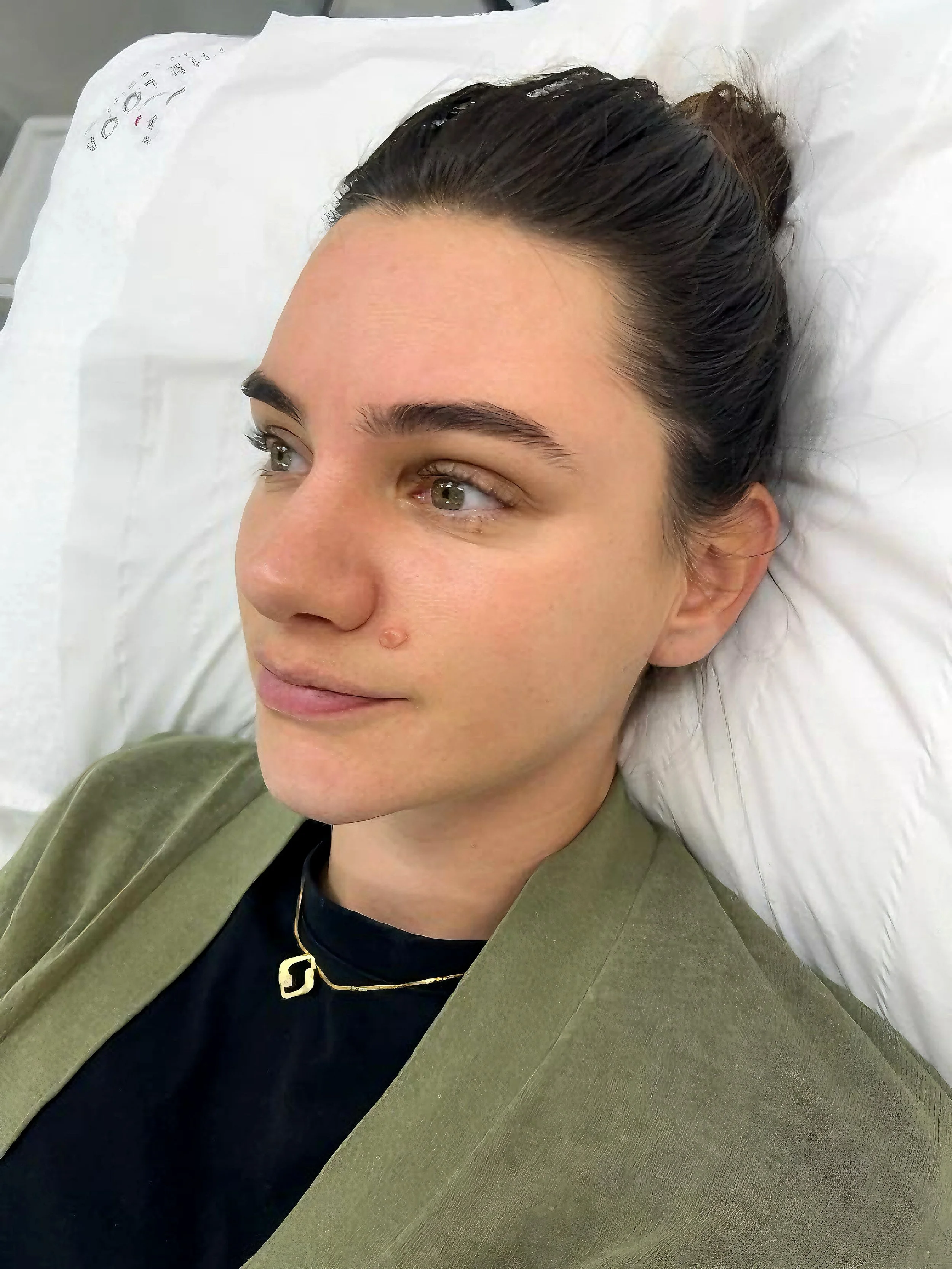 Close-up of a woman lying in a hospital bed, with dark hair and light skin, wearing a gold necklace with a geometric pendant and a green jacket.