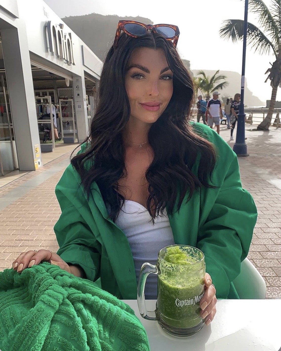 A woman with long dark wavy hair, sunglasses on her head, sitting outdoors at a cafe, holding a mug of green smoothie, with a beach, palm trees, and people in the background.