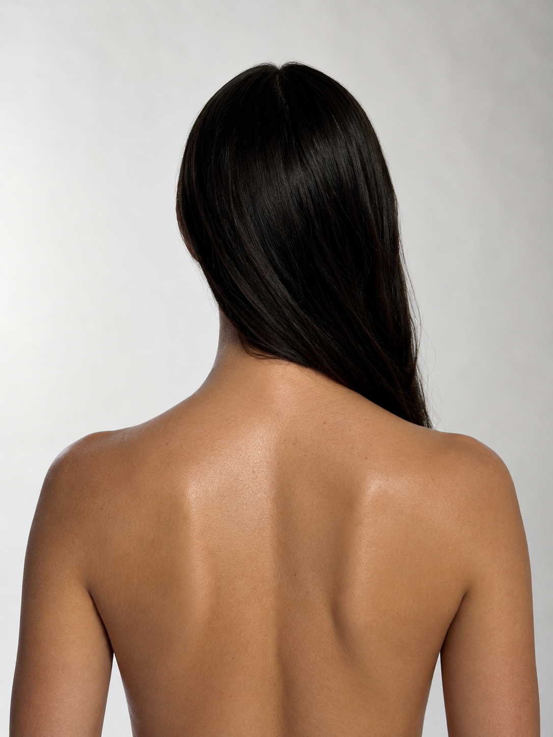 Back of a woman with long, straight, dark hair and smooth, tan skin, seen from behind against a plain light background.