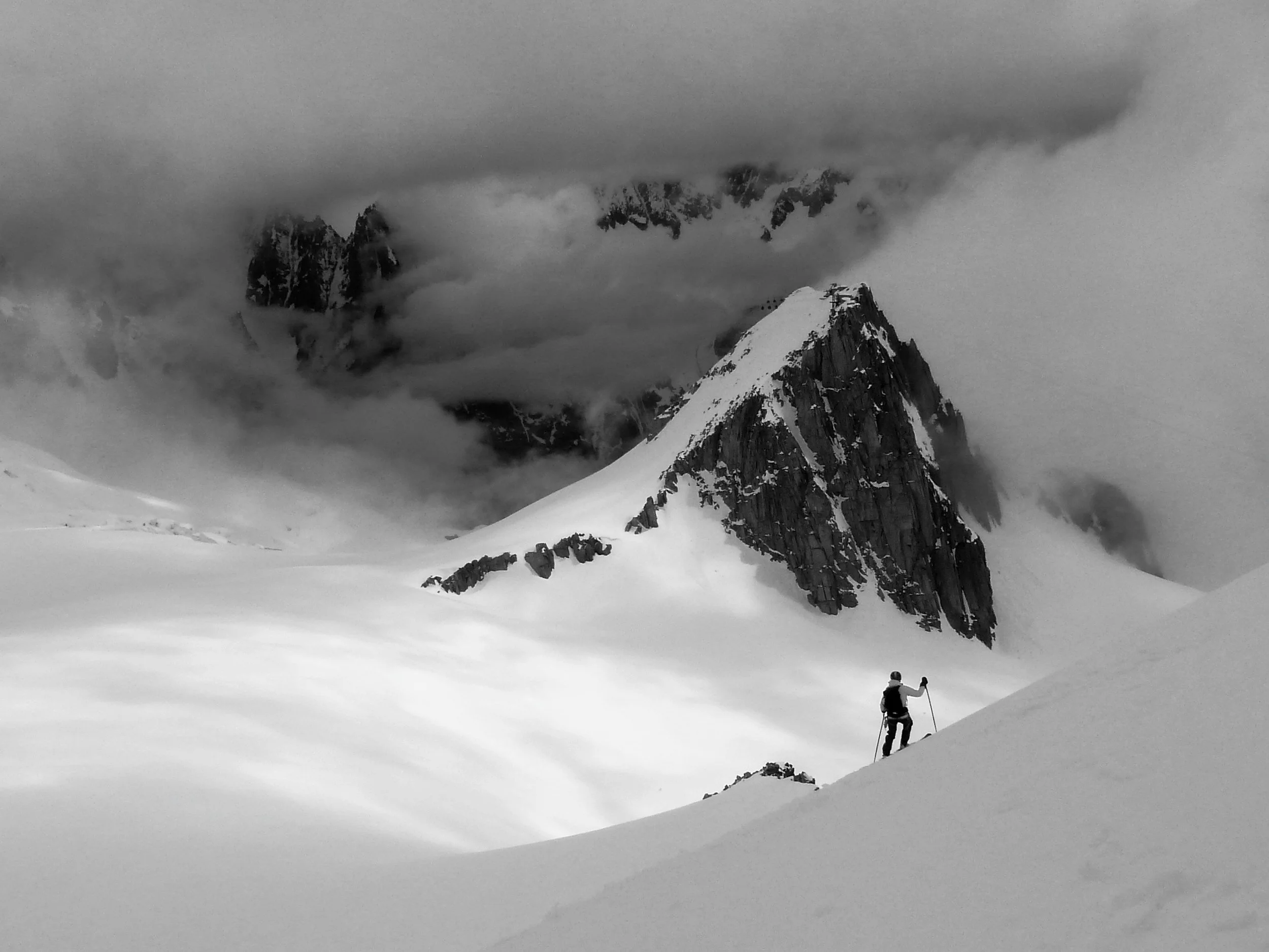 A person hiking in snowy mountain terrain, with towering snow-covered peaks in the background partially obscured by clouds.