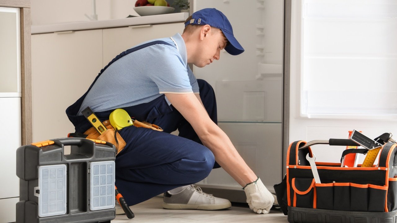 A male handyman wearing a blue cap, light blue shirt, and work gloves kneeling on the floor, using a tool near an open toolbox in a bright kitchen.