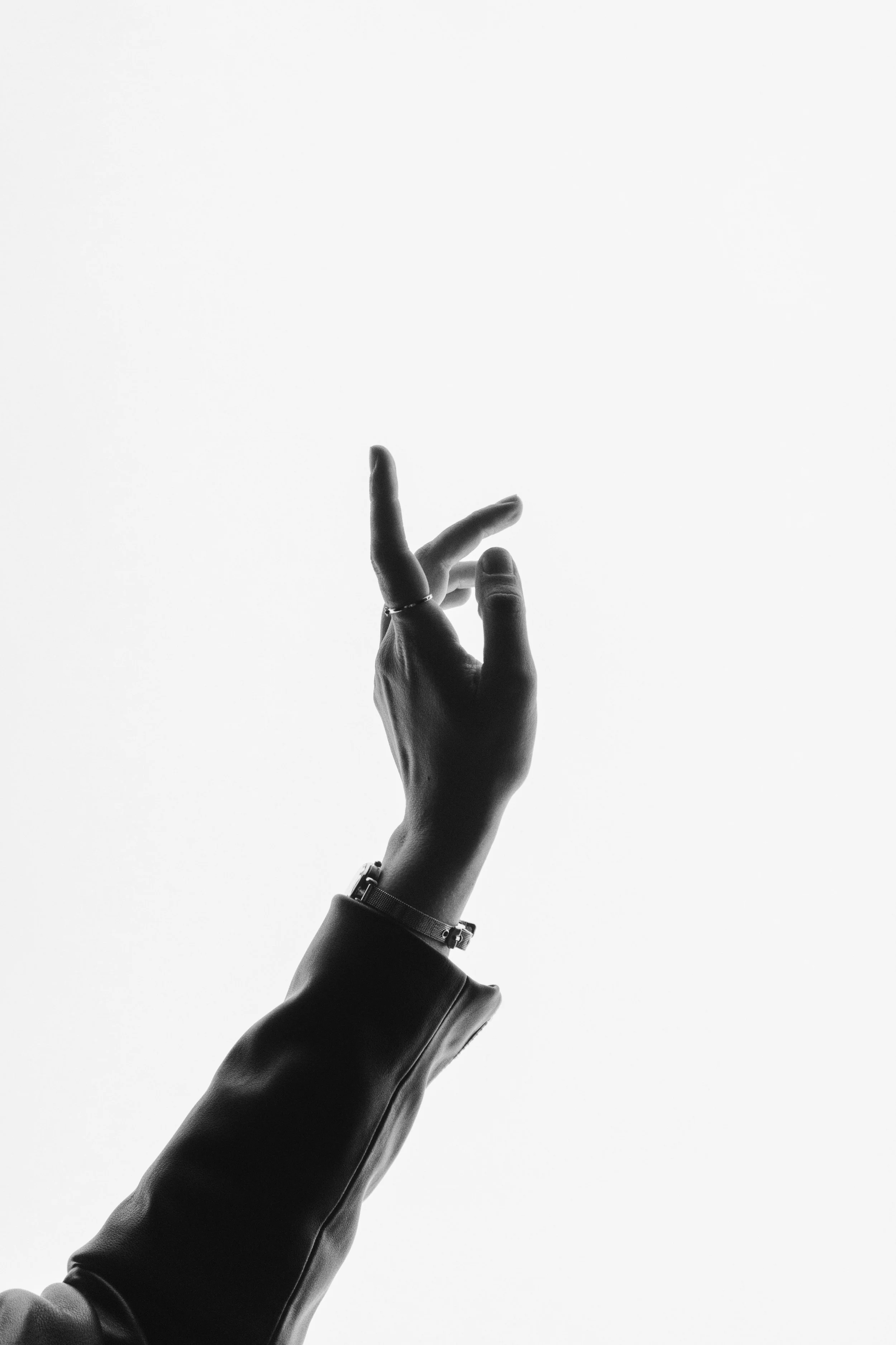 Black and white photo of a hand with a ring and bracelet, pointing or reaching upward against a plain background.
