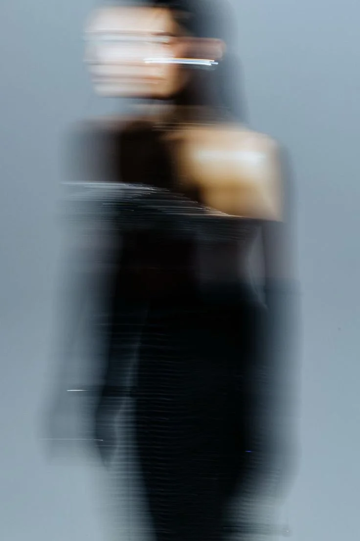Blurred portrait of a woman wearing glasses and a black off-shoulder top, with dark hair and a gray background.