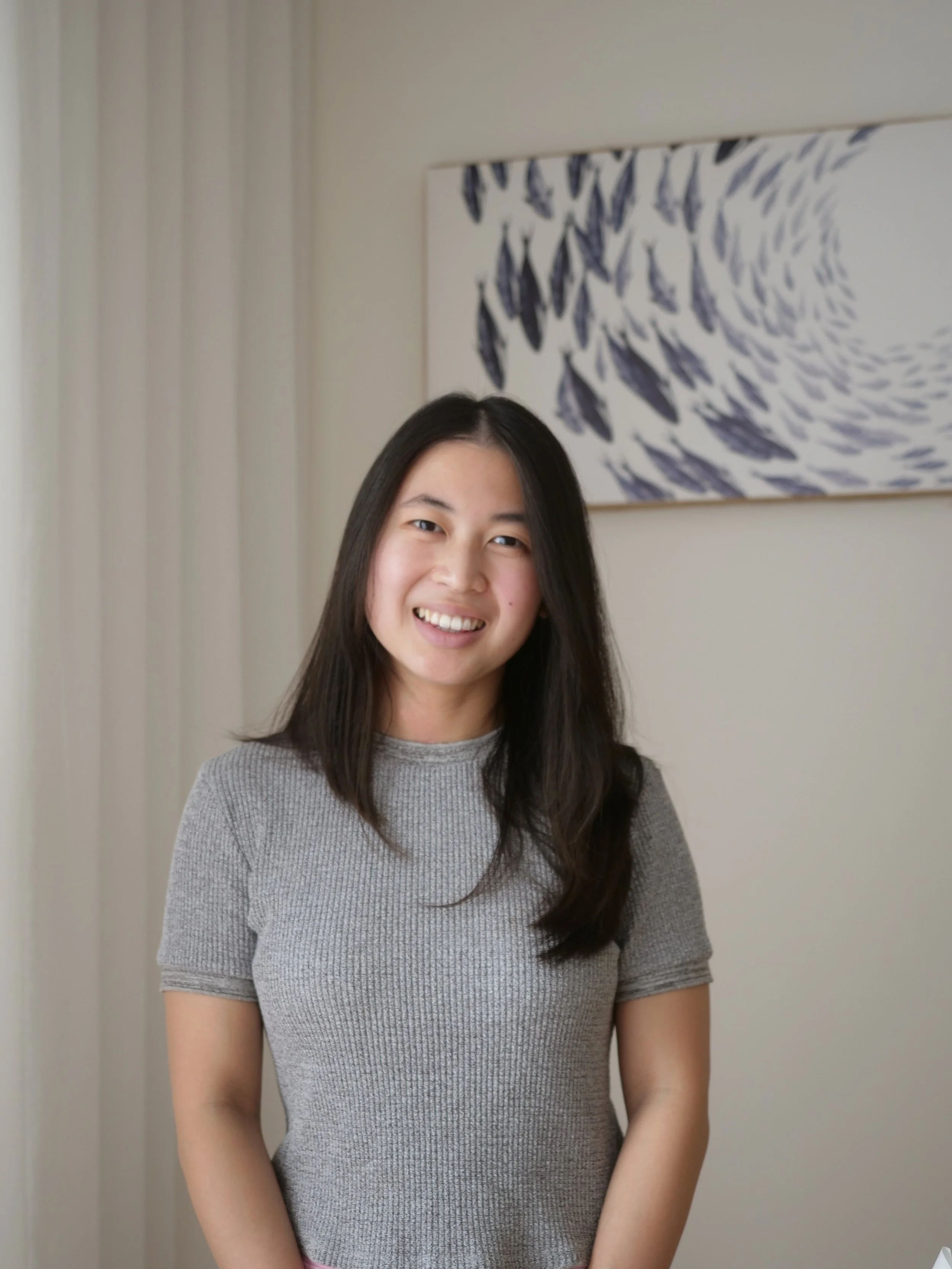 A woman with long dark hair, wearing a grey T-shirt, smiling, standing in front of a white wall with a large, abstract, blue and white painting. There is a white curtain on the left side of the image.